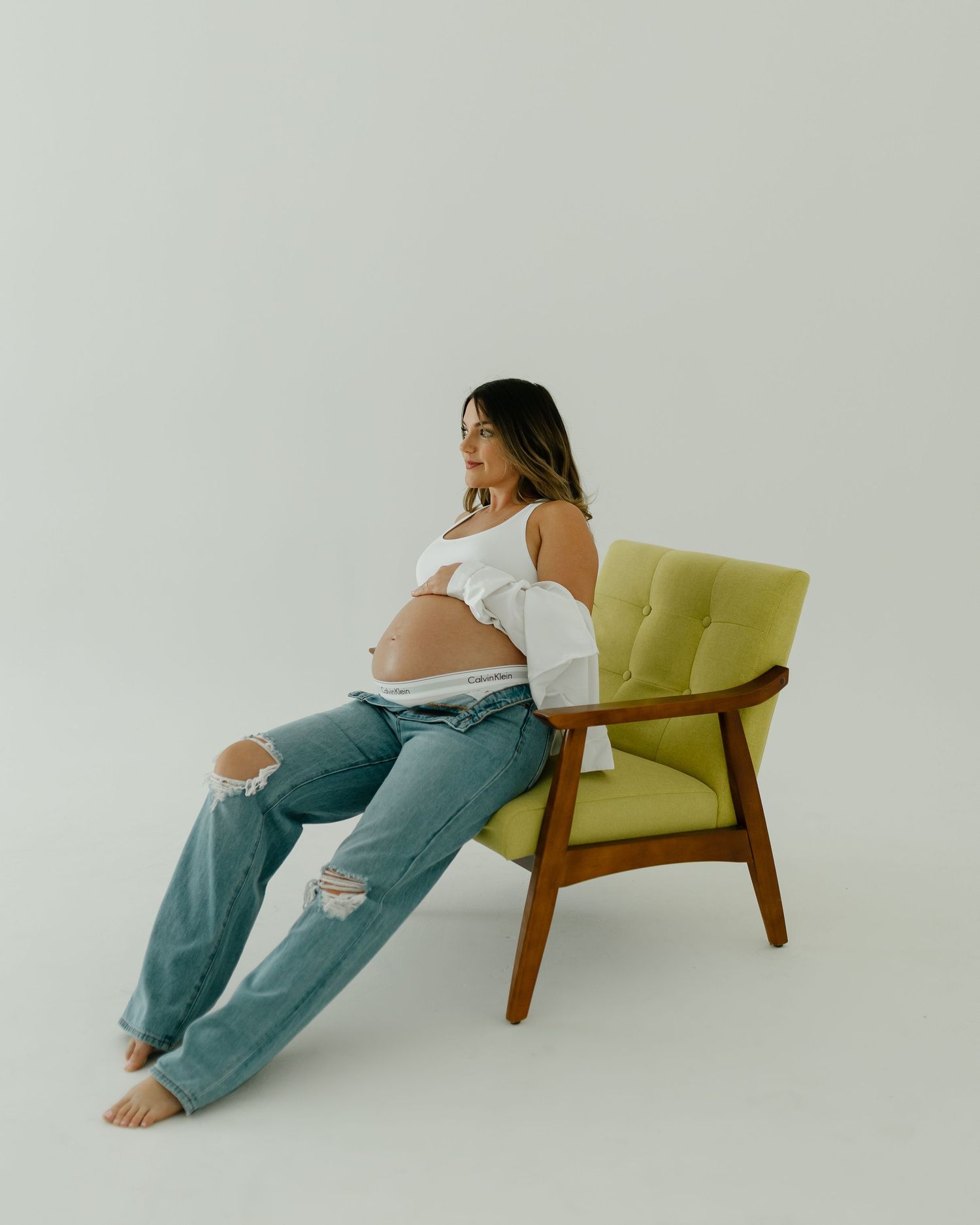 Pregnant woman in white top and ripped jeans sitting on a green mid-century modern chair in a studio.