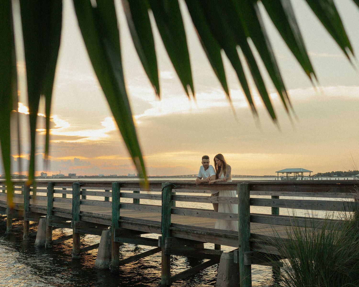 Couple standing together on a waterfront pier at golden sunset framed by palm leaves.