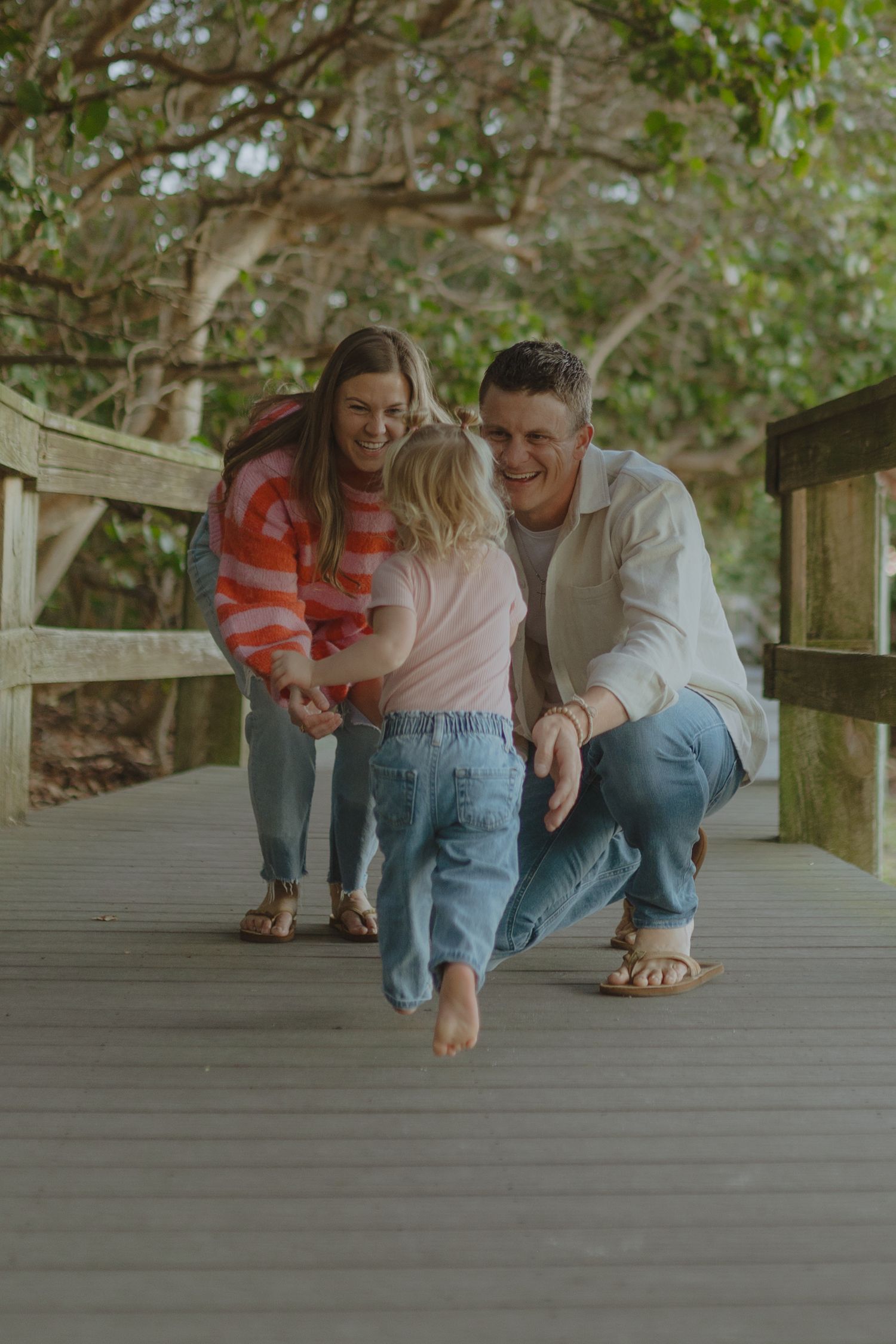 A joyful family moment on a wooden bridge, parents laughing as their toddler takes steps toward dad outdoors.