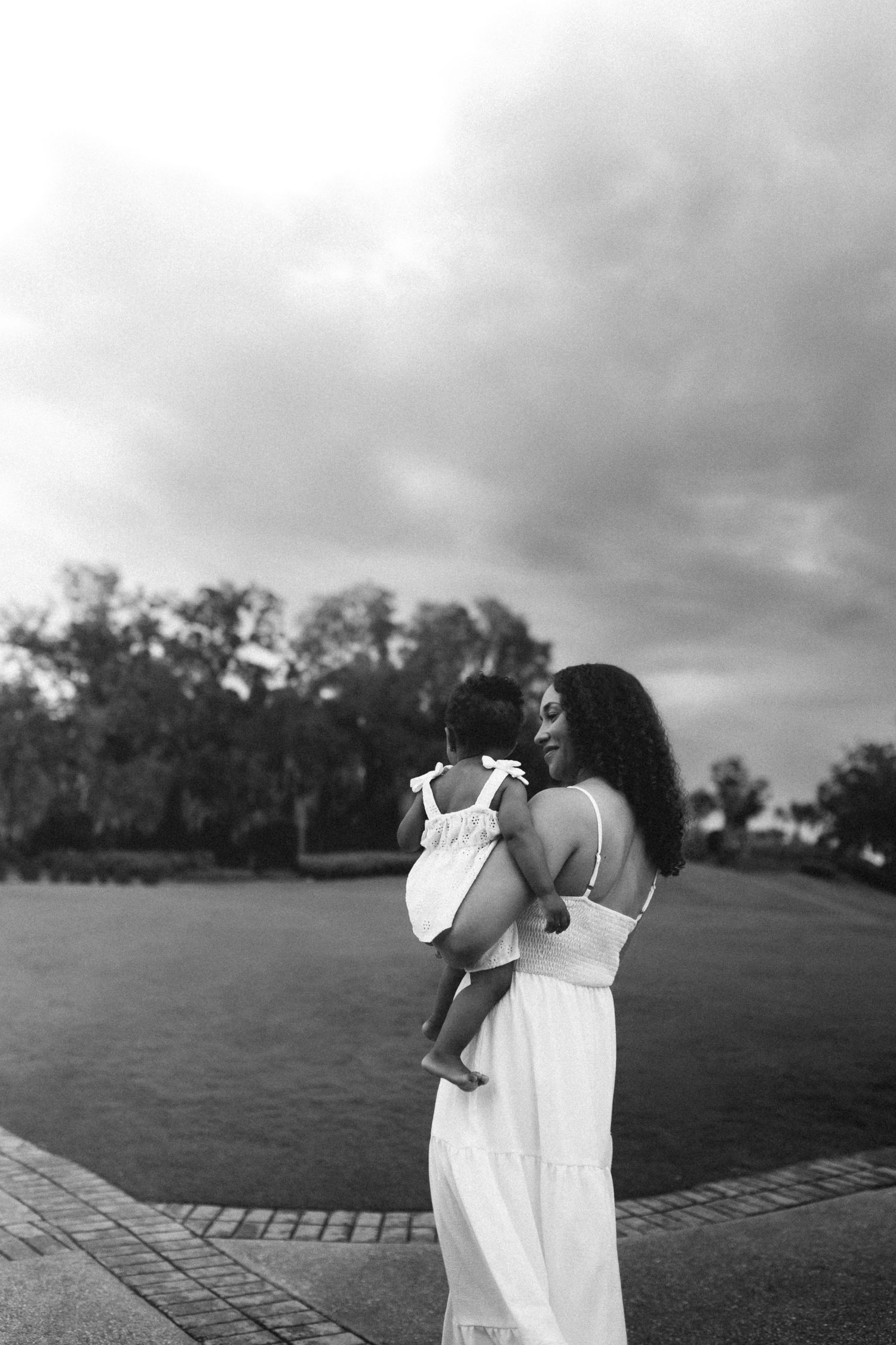 Black and white photo of a mother in white dress tenderly holding her baby outdoors under cloudy sky.