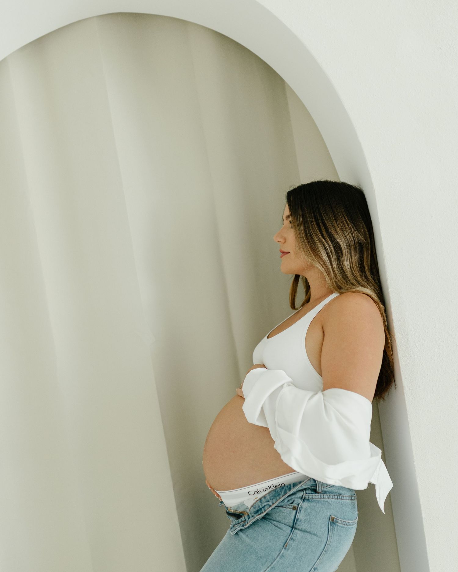 Pregnant woman in white crop top and jeans standing in an arched doorway cradling her baby bump.