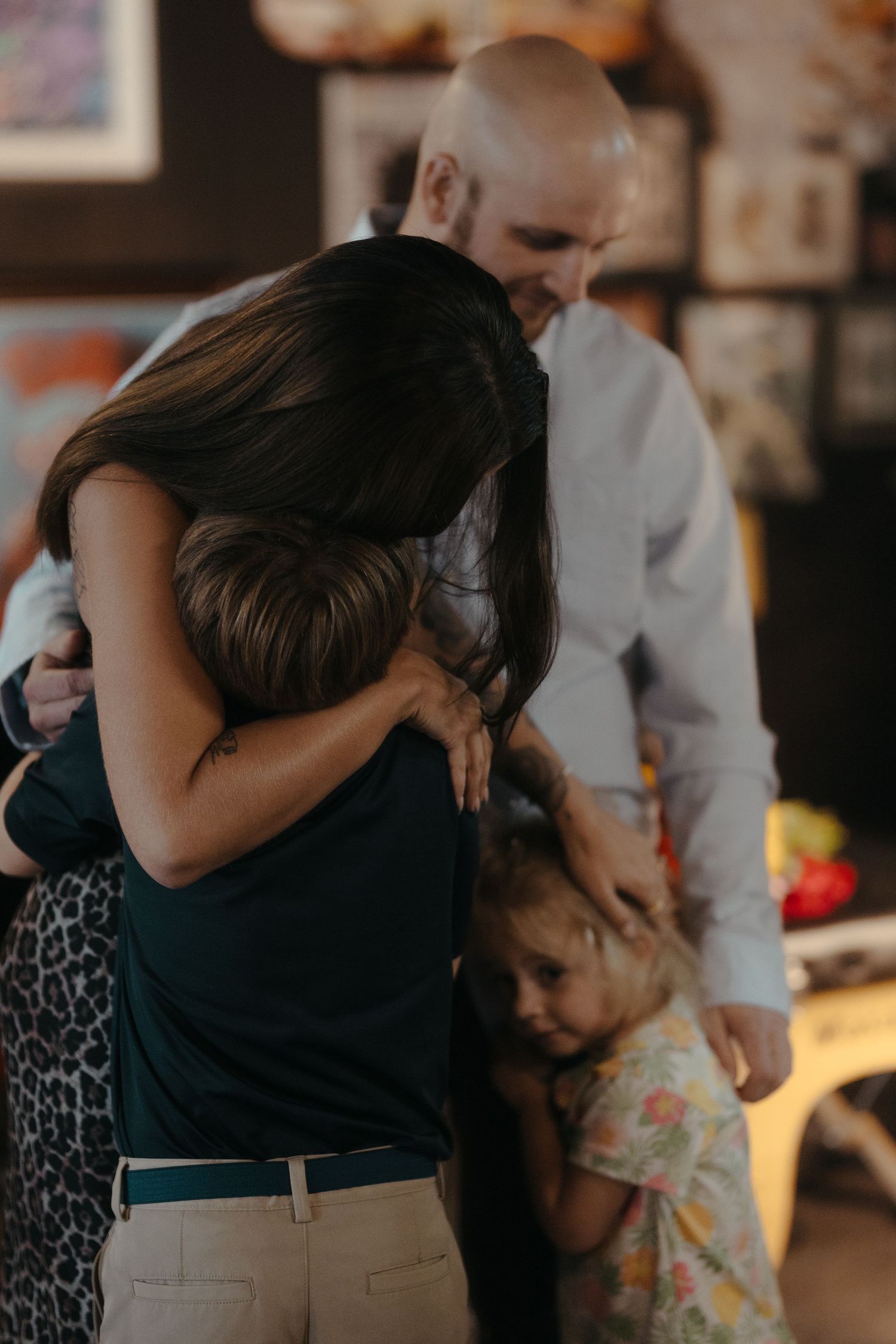 A tattooed woman hugs a child while a bald man smiles warmly in a cozy indoor setting.