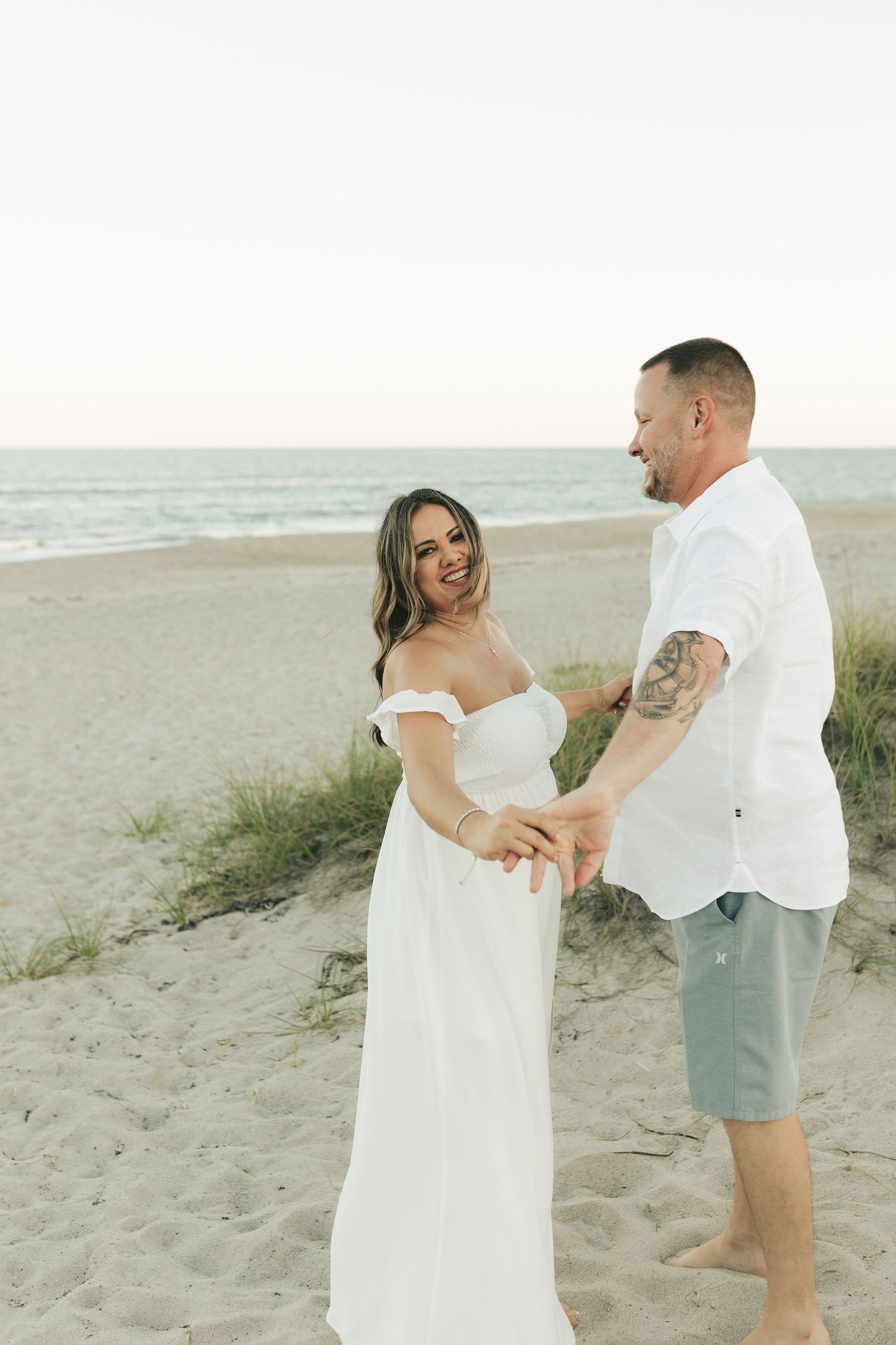Couple holding hands on a sandy beach at sunset, woman in white off-shoulder gown, man in white shirt and blue shorts.