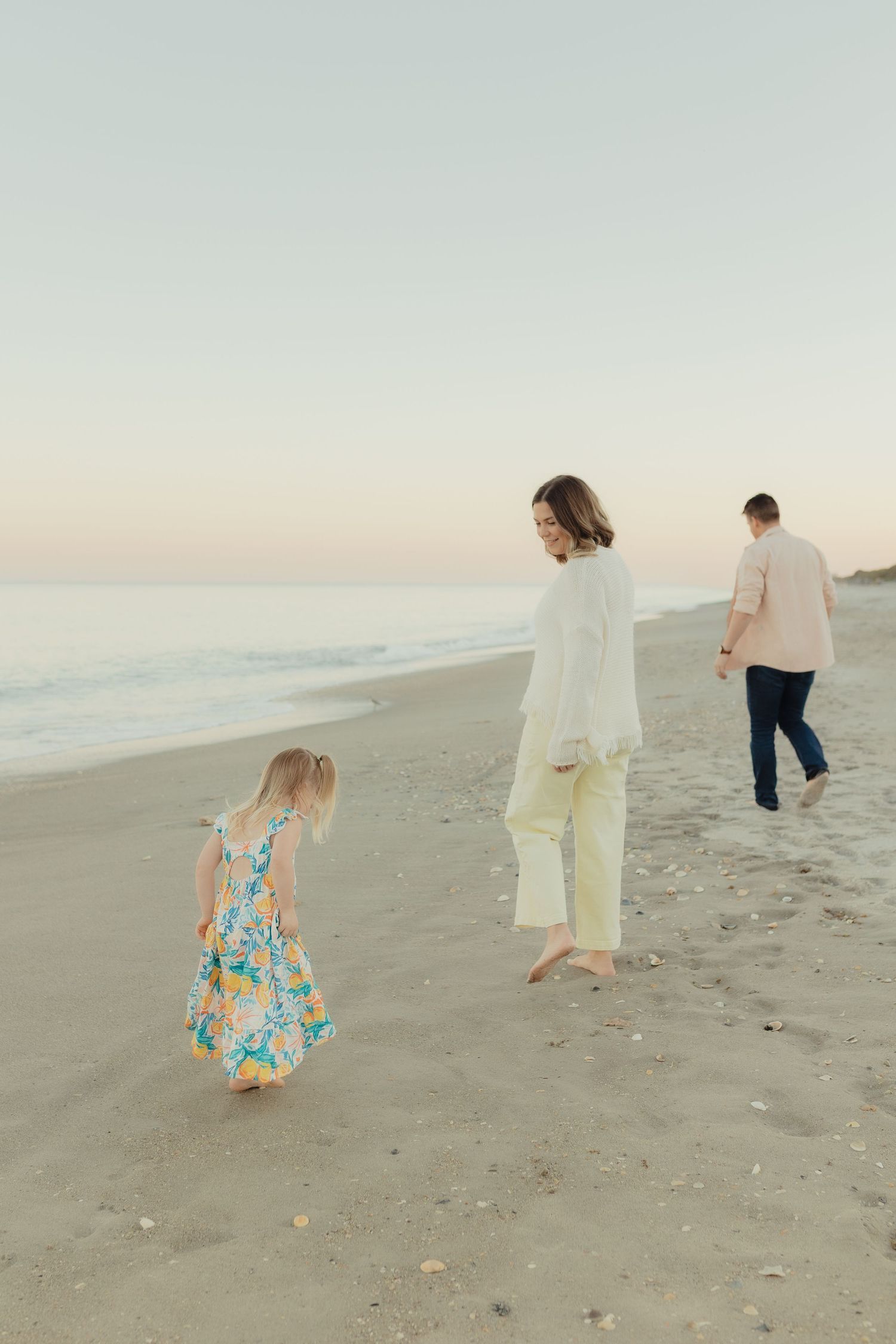 Family of three walking on a sandy beach at dusk, mother in white top looks back at young girl in floral dress.