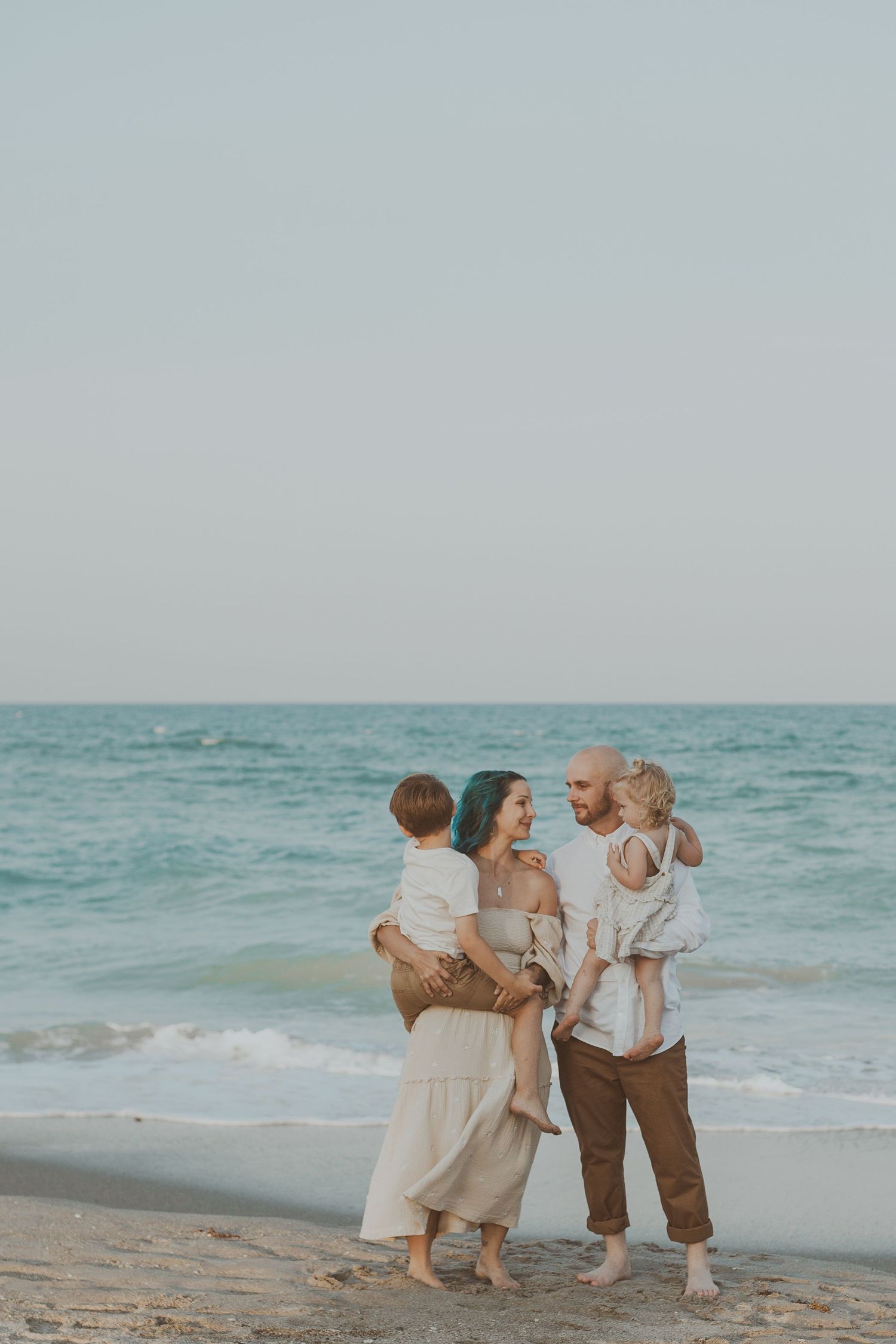 Family of four posing on a sandy beach at sunset, parents holding toddlers in neutral tones with ocean waves behind.