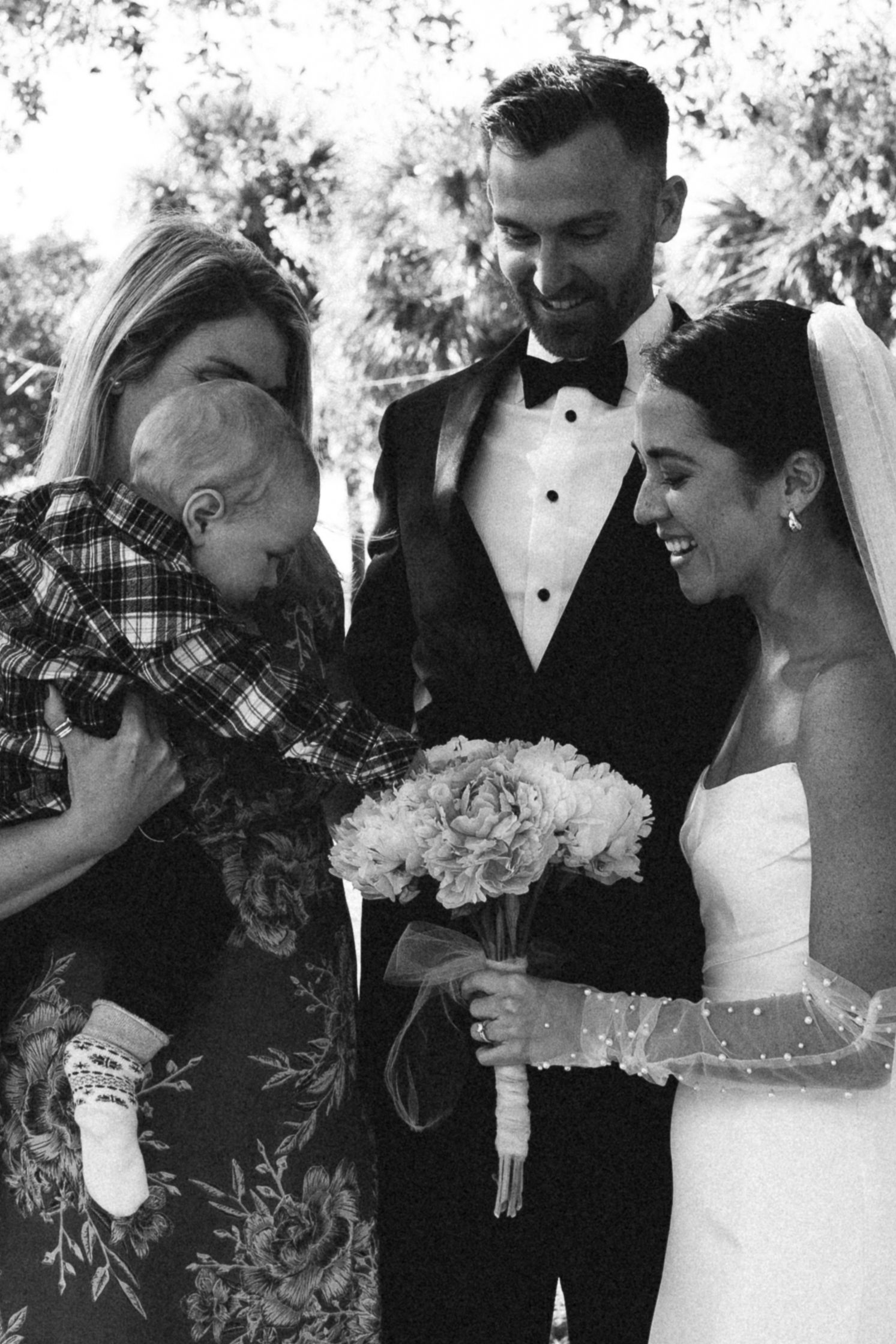 Black and white wedding photo of bride and groom smiling with woman holding baby at outdoor ceremony.