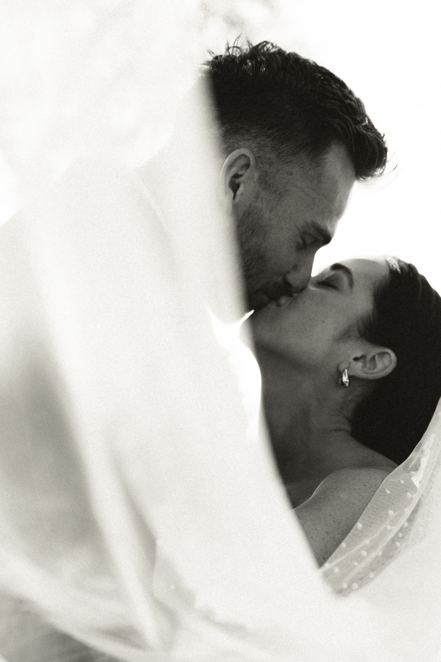 Romantic black and white photo of bride and groom kissing beneath flowing wedding veil.