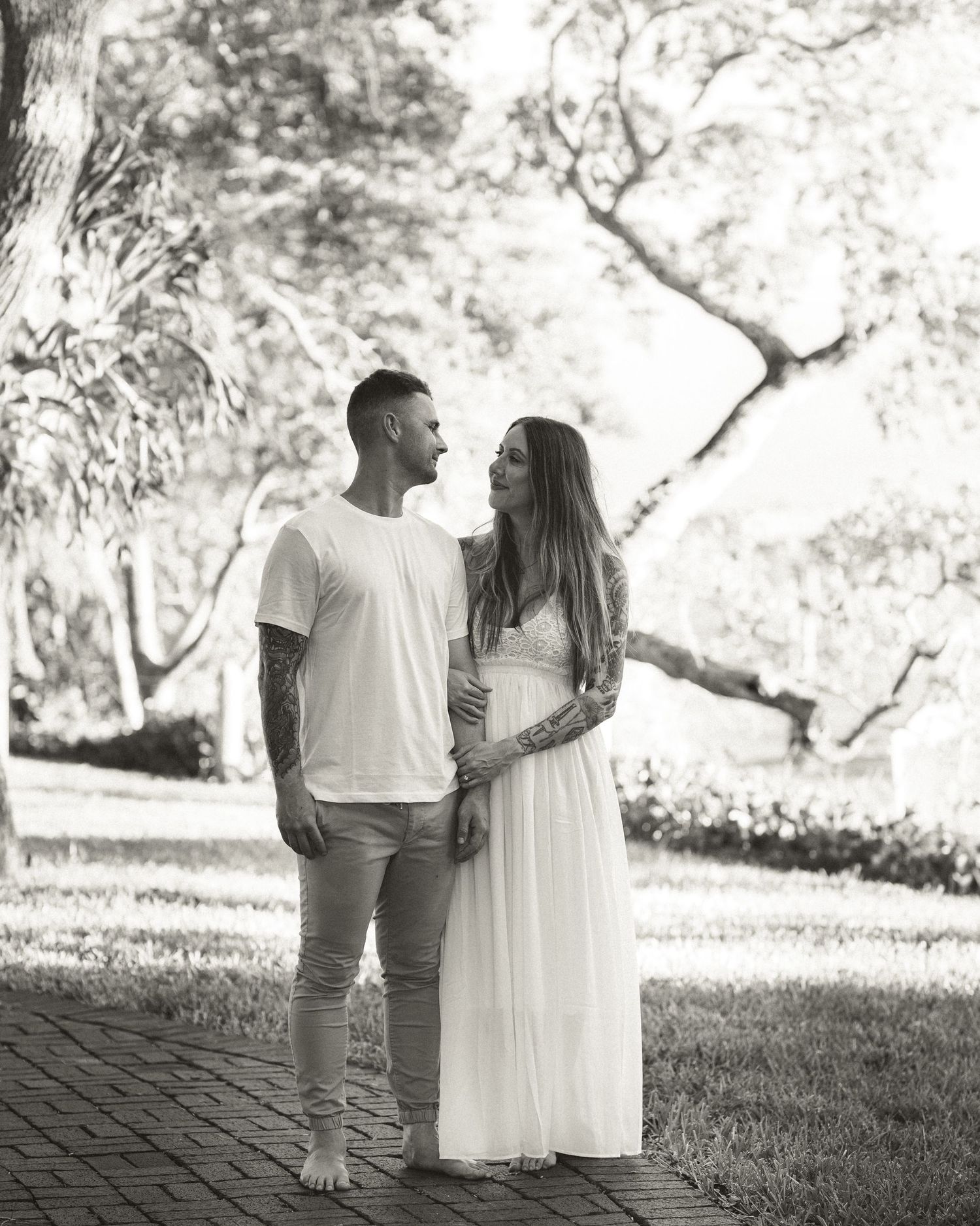 Couple gazing at each other on a brick path in a park, woman in white maternity dress, black and white photo.