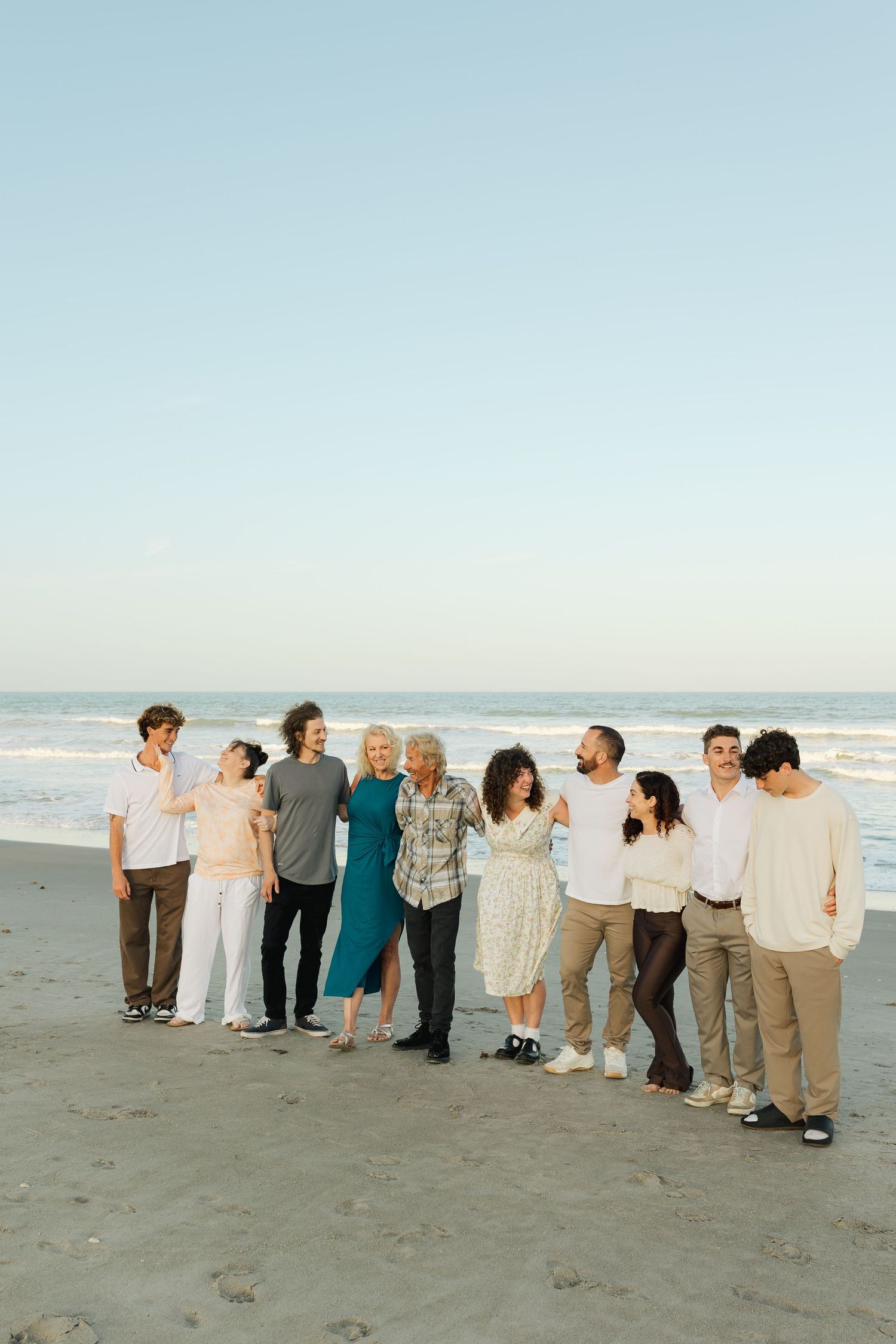 Large family group posing together on a sandy beach at sunset, smiling and embracing in casual neutral outfits.