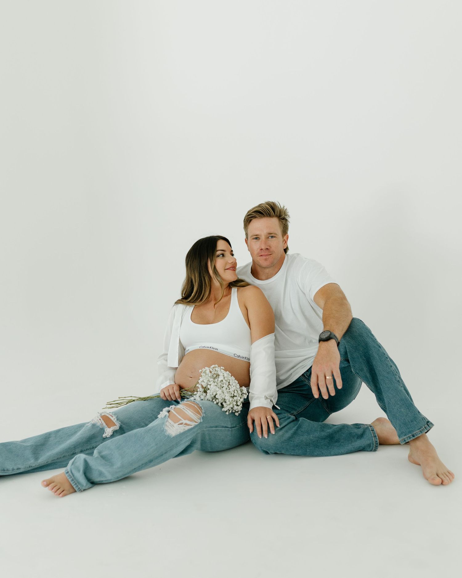 Couple sitting on floor in white tops and ripped jeans for a casual maternity photo with baby breath flowers.