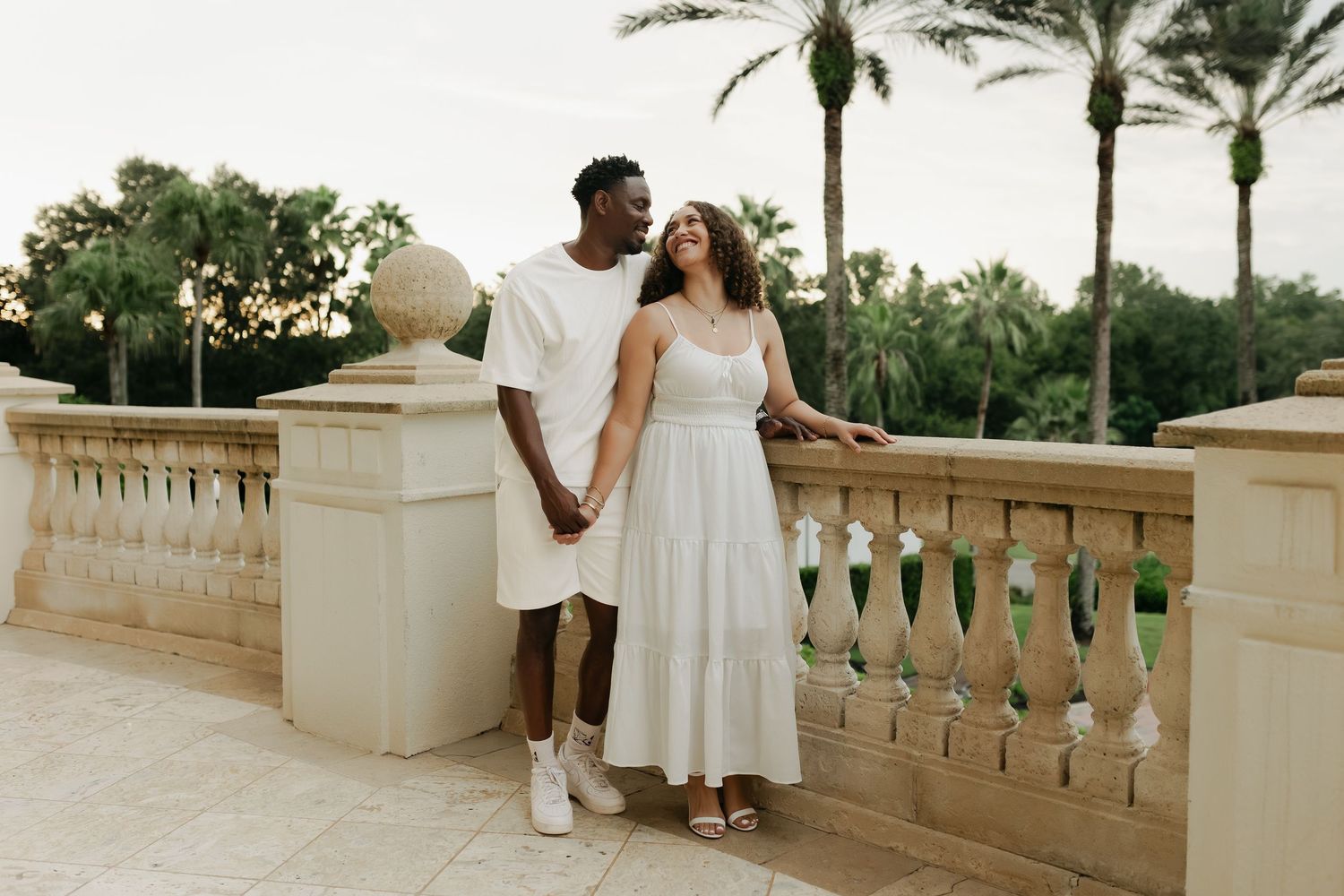 Young couple in white outfits holding hands on a terrace with palm trees at dusk.