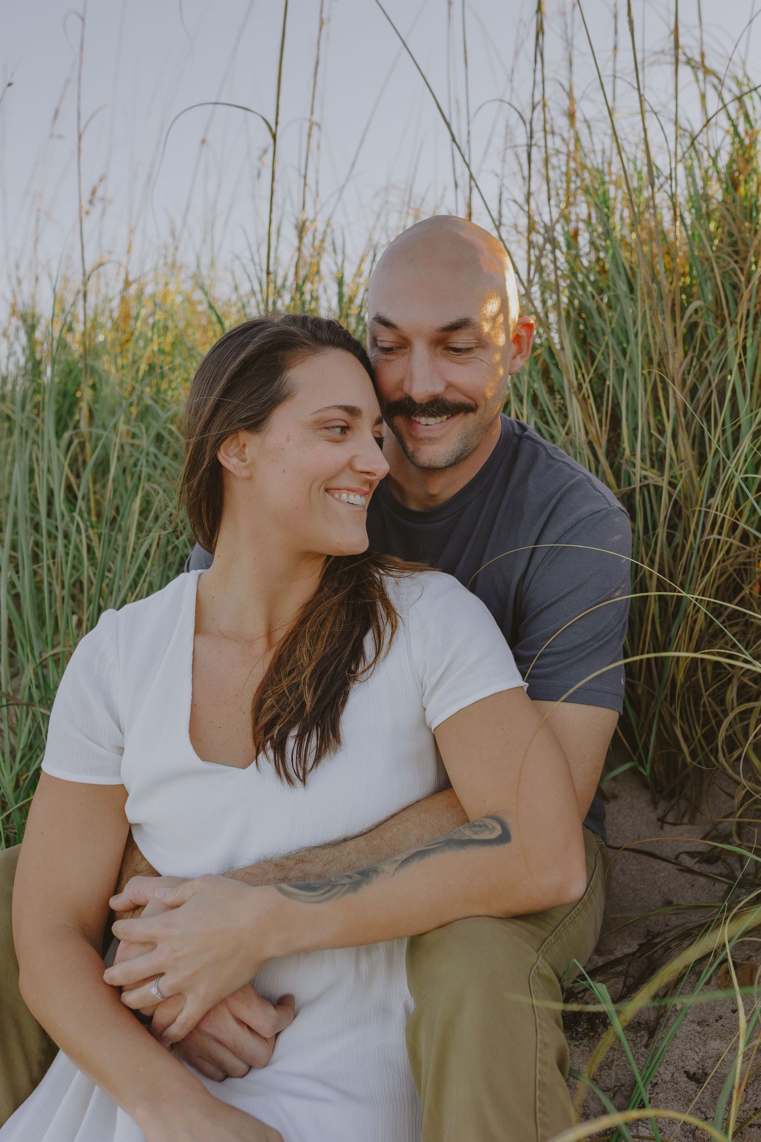 Couple sitting together in tall beach grass, man with mustache embracing smiling woman in white top.