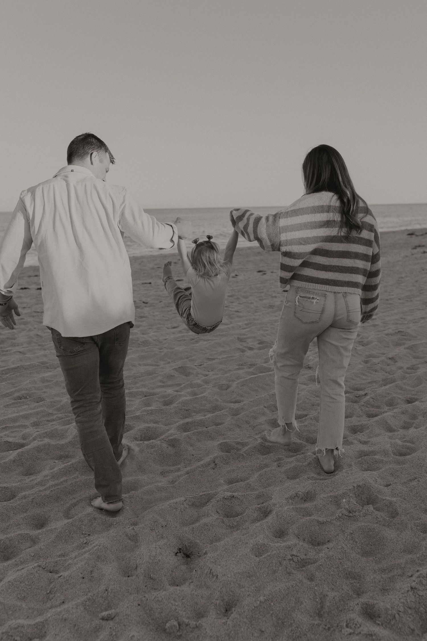 Black and white photo of parents swinging toddler by hands while walking on sandy beach.
