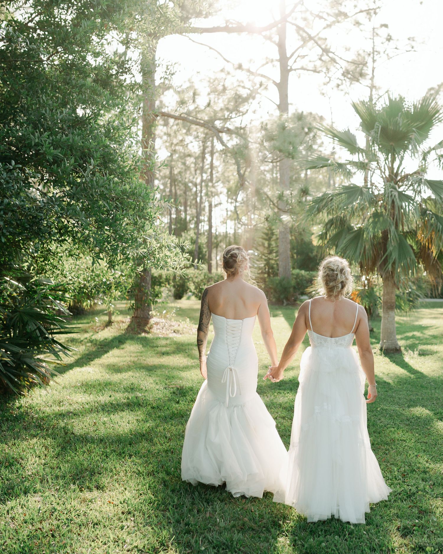 Two brides in white wedding gowns hold hands walking through a scenic tree-lined lawn.