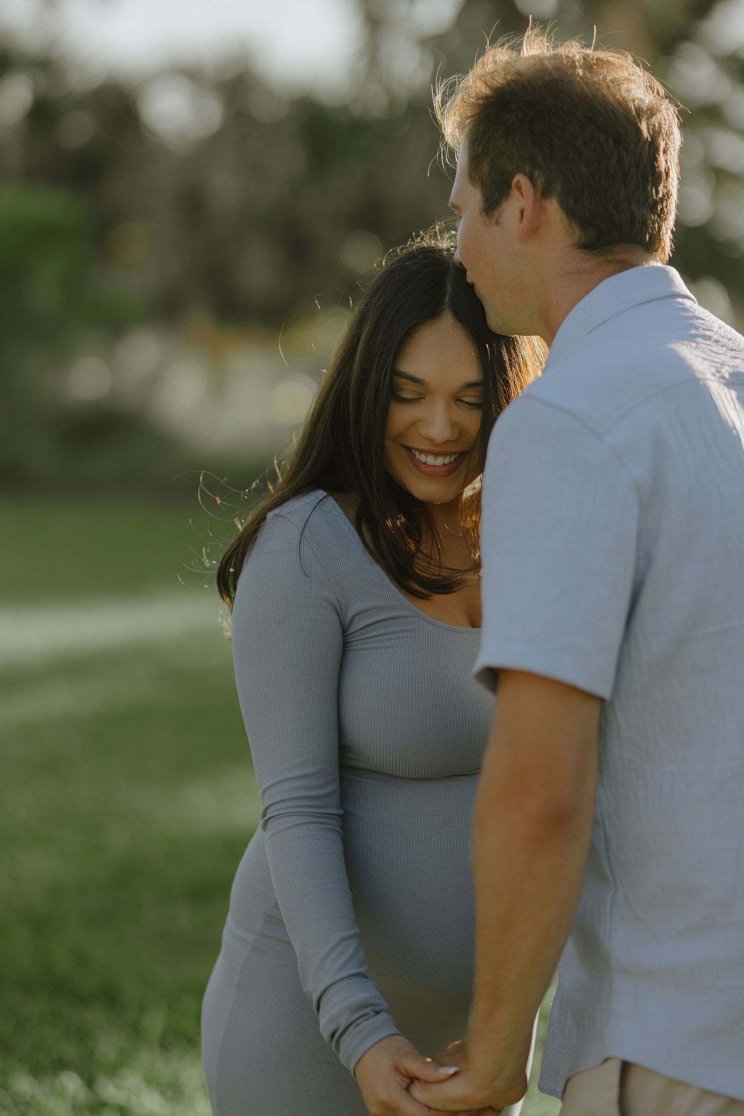 Smiling pregnant woman in a grey dress holding hands with her partner in a sunlit outdoor setting.