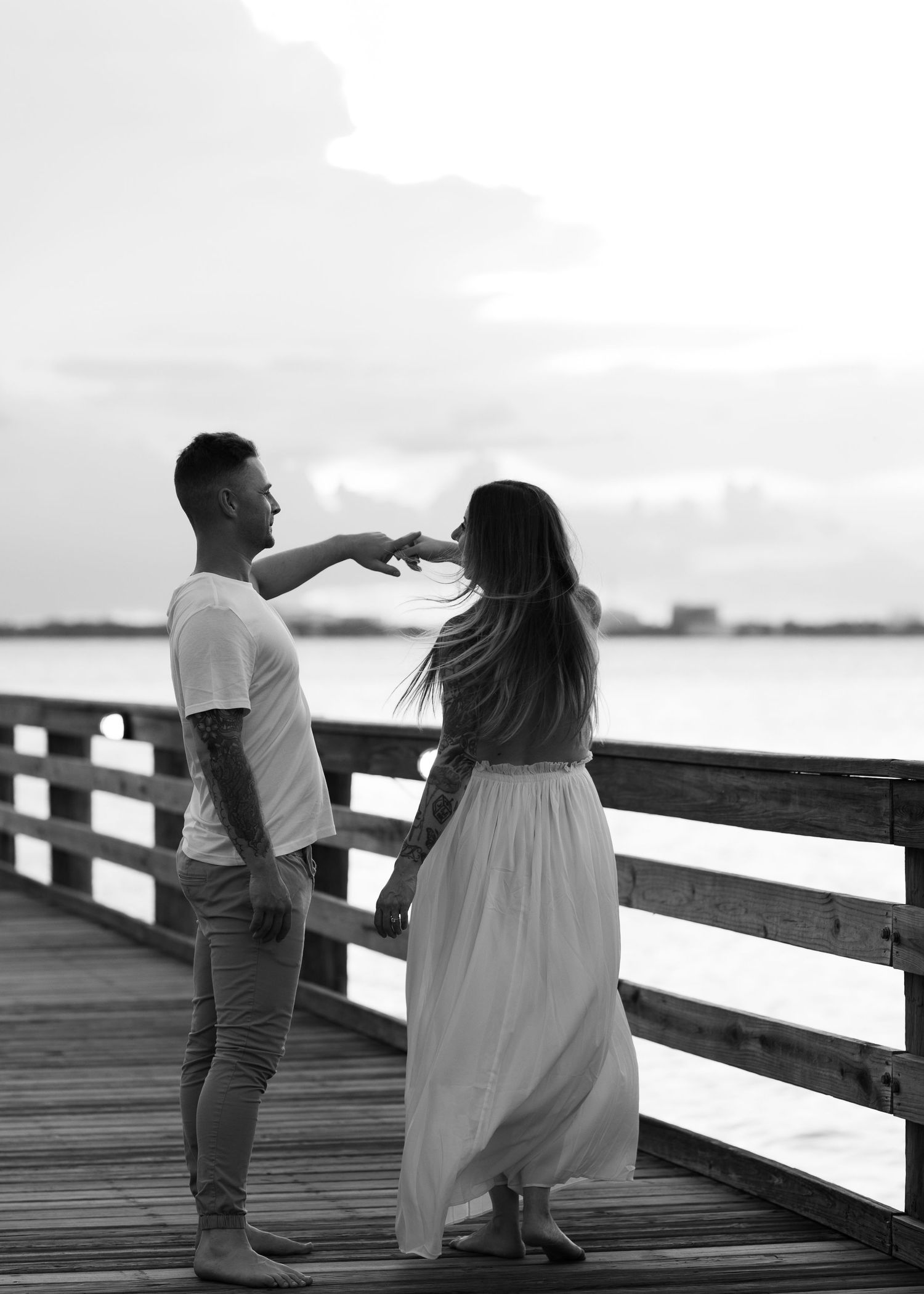 Black and white photo of a tattooed couple holding hands on a waterfront pier at sunset.