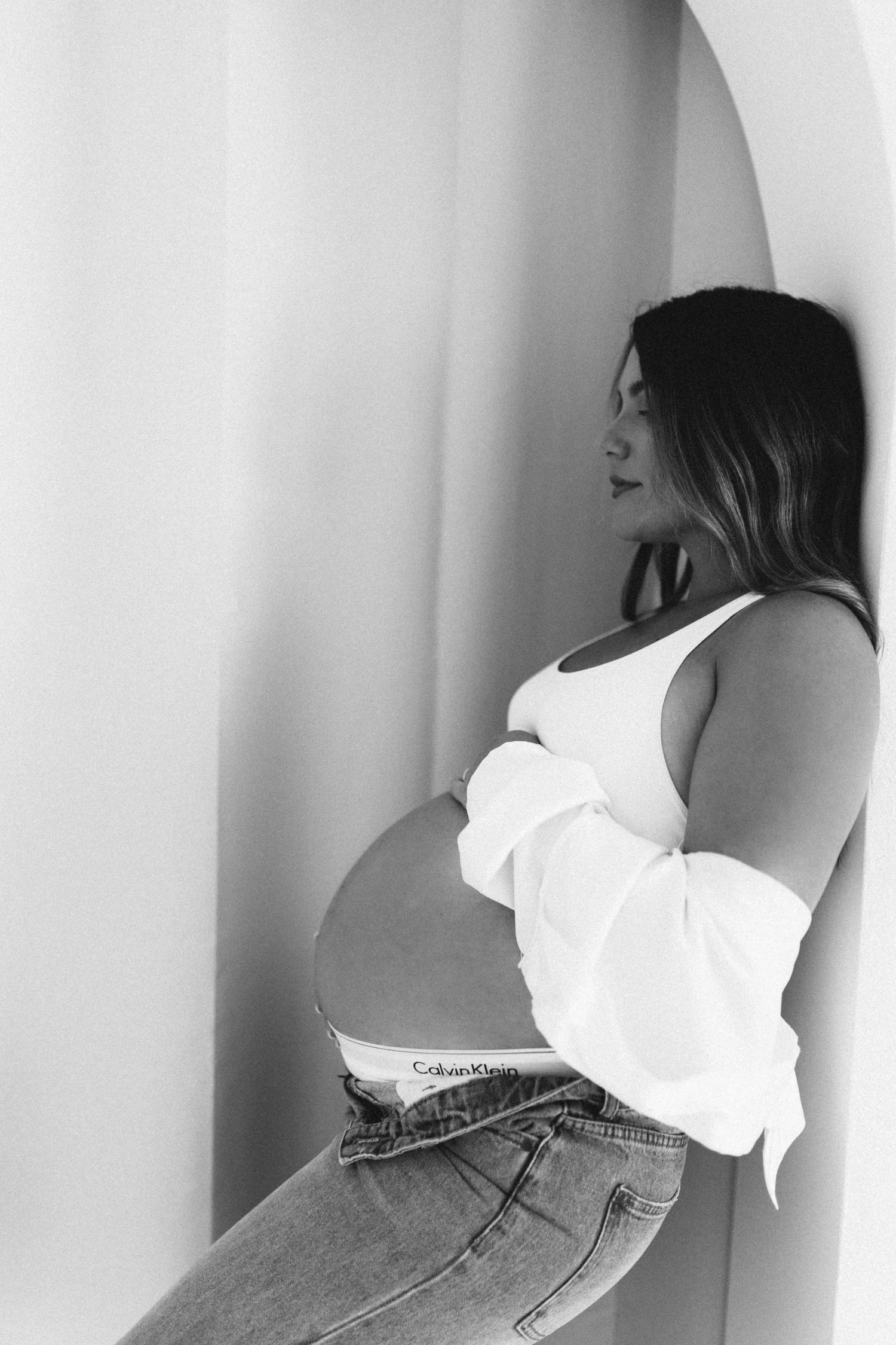 Black and white photo of a pregnant woman in jeans and white top leaning against an arched wall.