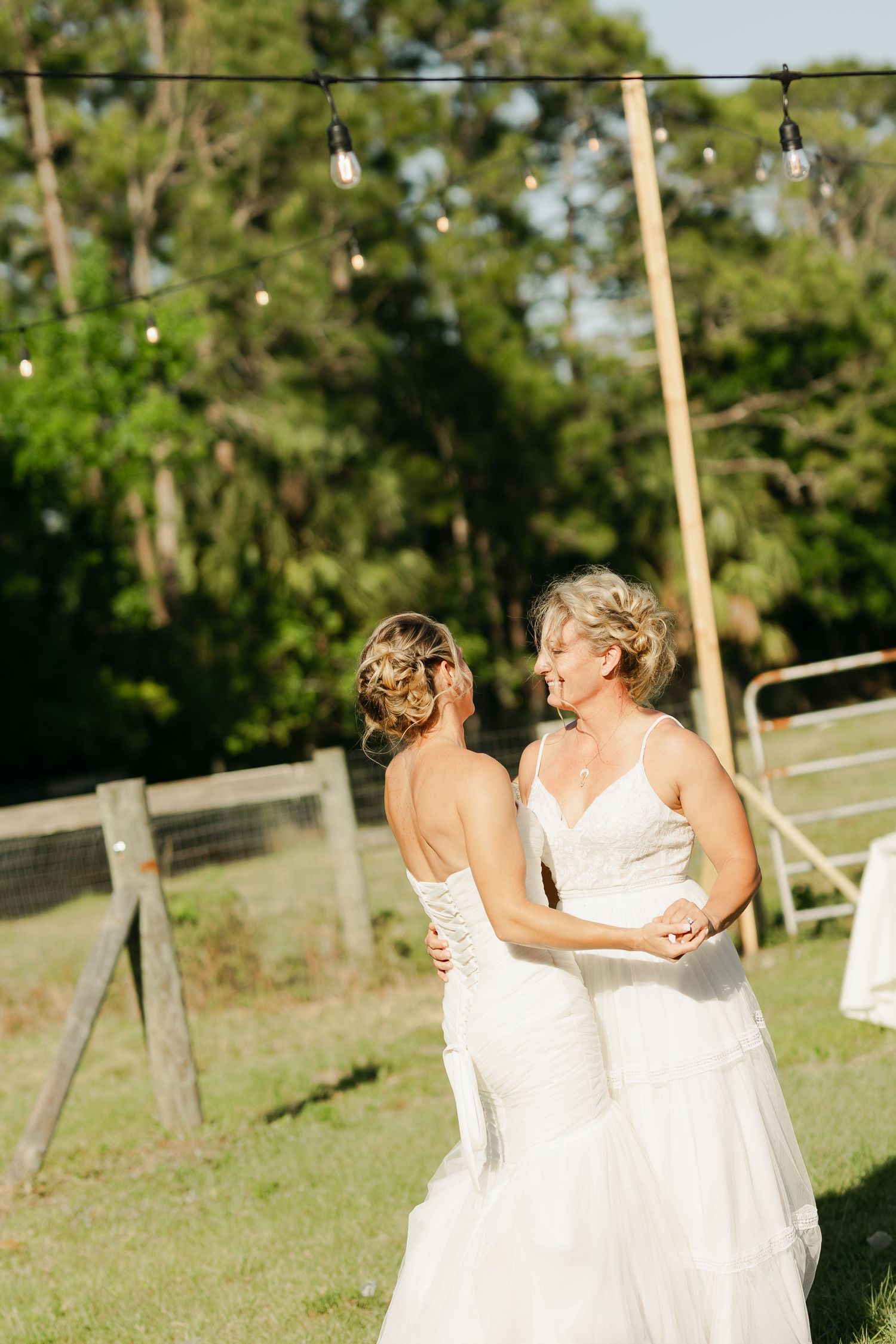 Two brides in white wedding dresses share a first dance outdoors under string lights at sunset.