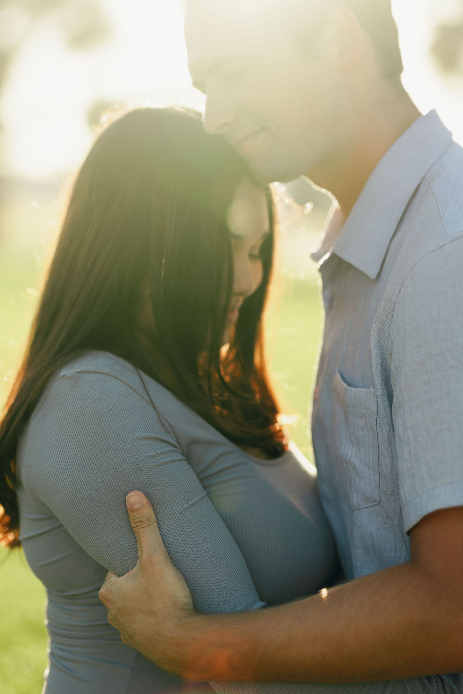Pregnant couple embracing in golden sunlight, man holding woman's baby bump in a tender outdoor moment.