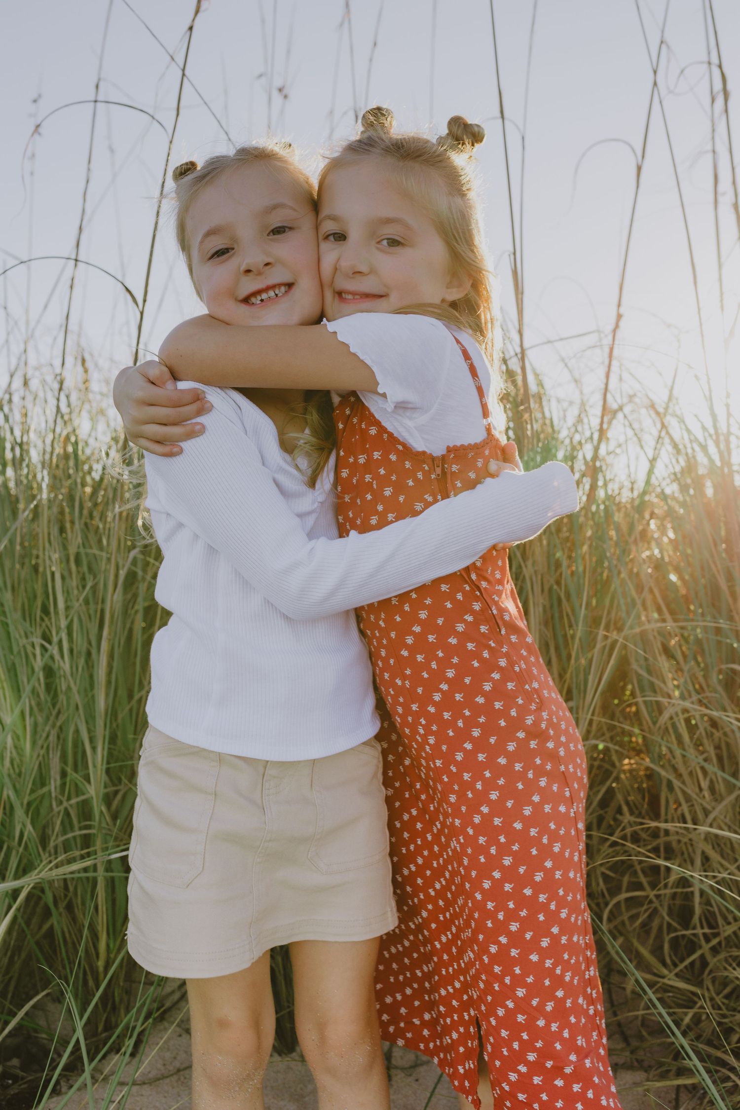 Two young girls with matching bun hairstyles hugging each other outdoors near tall grass in golden sunlight.