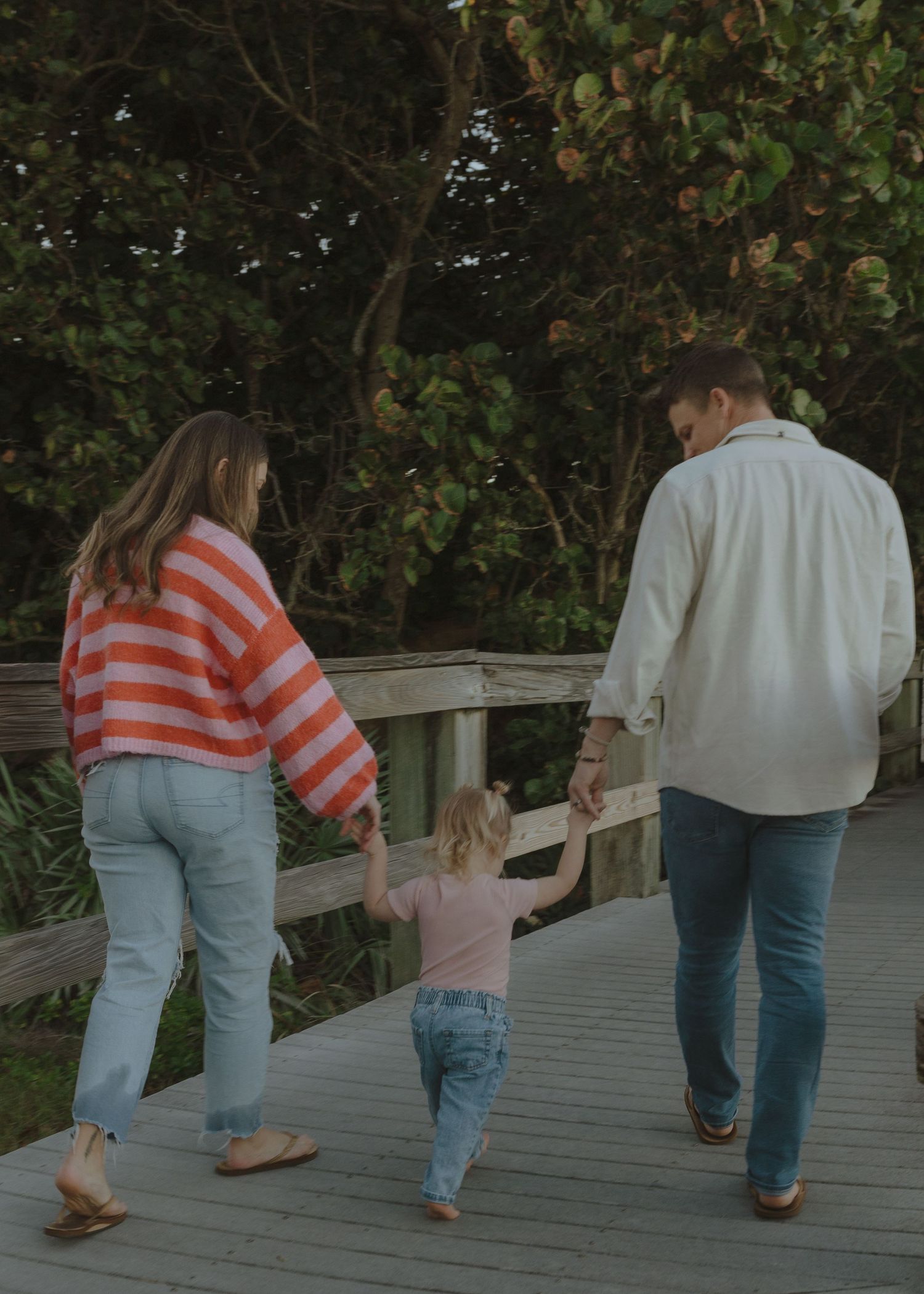 A family of three walks hand-in-hand on a wooden boardwalk surrounded by lush green trees.
