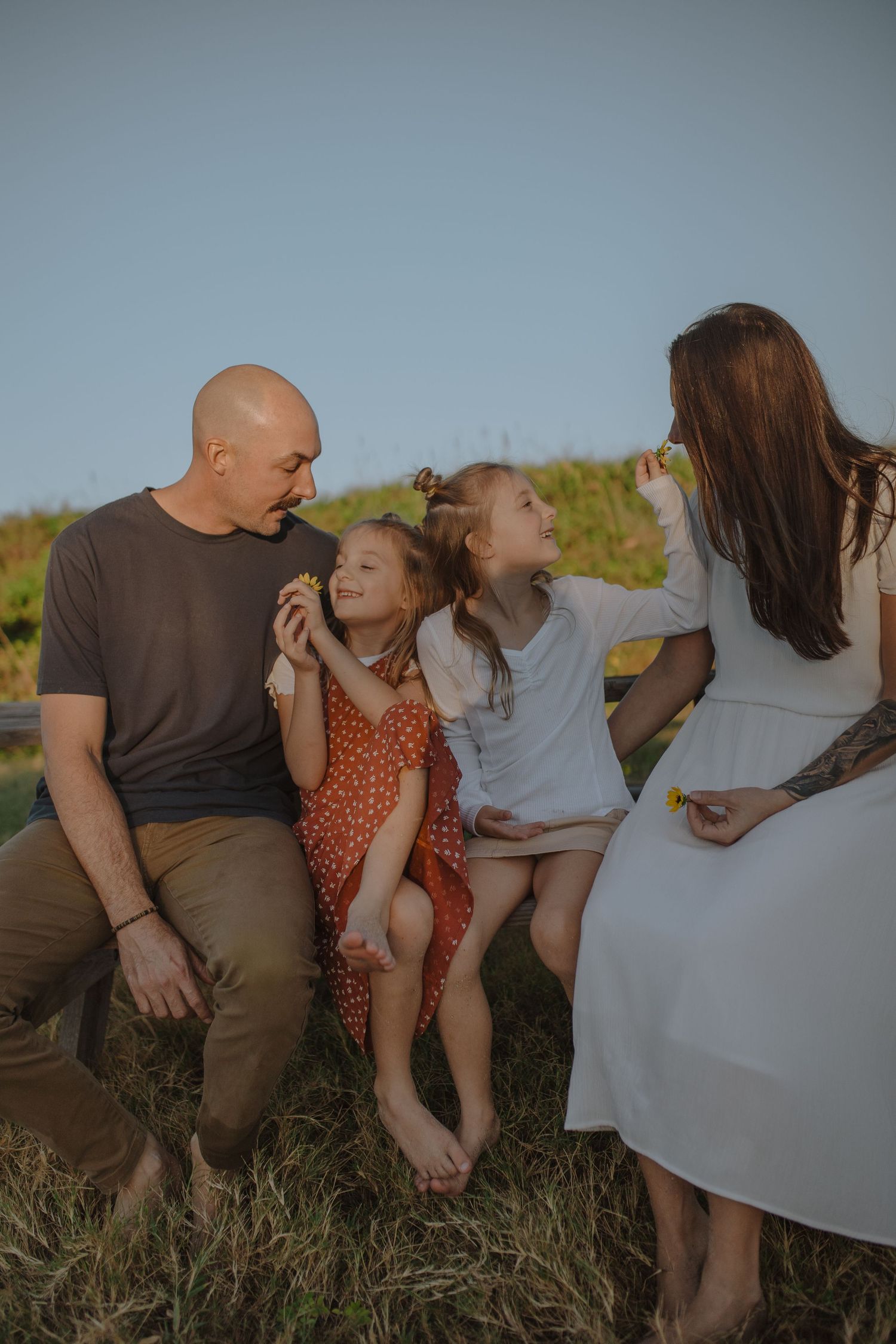 Happy family of four sitting on bench outdoors in golden hour light, parents smiling at two daughters.