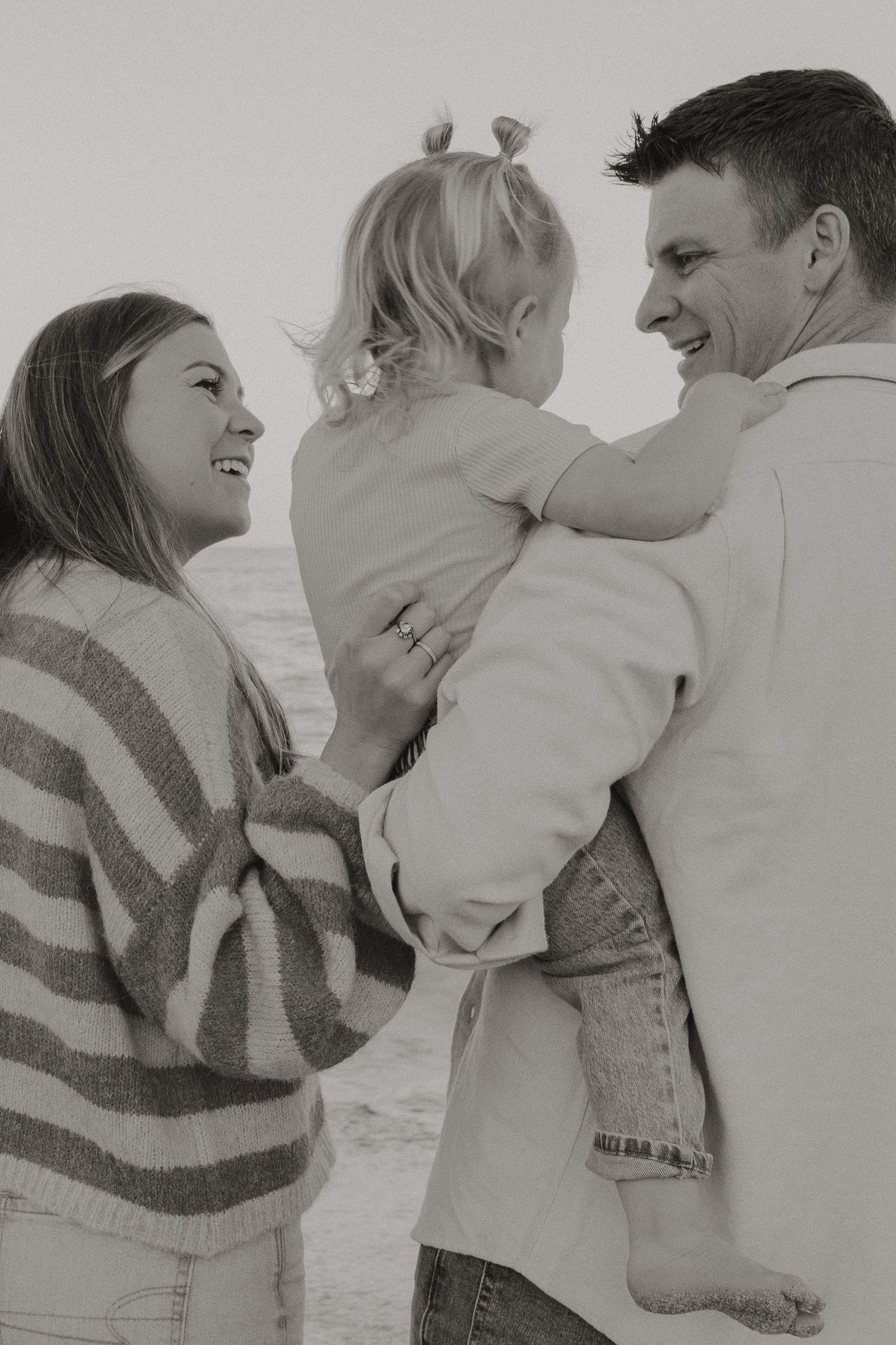 Black and white photo of parents smiling at toddler being held by father on beach.
