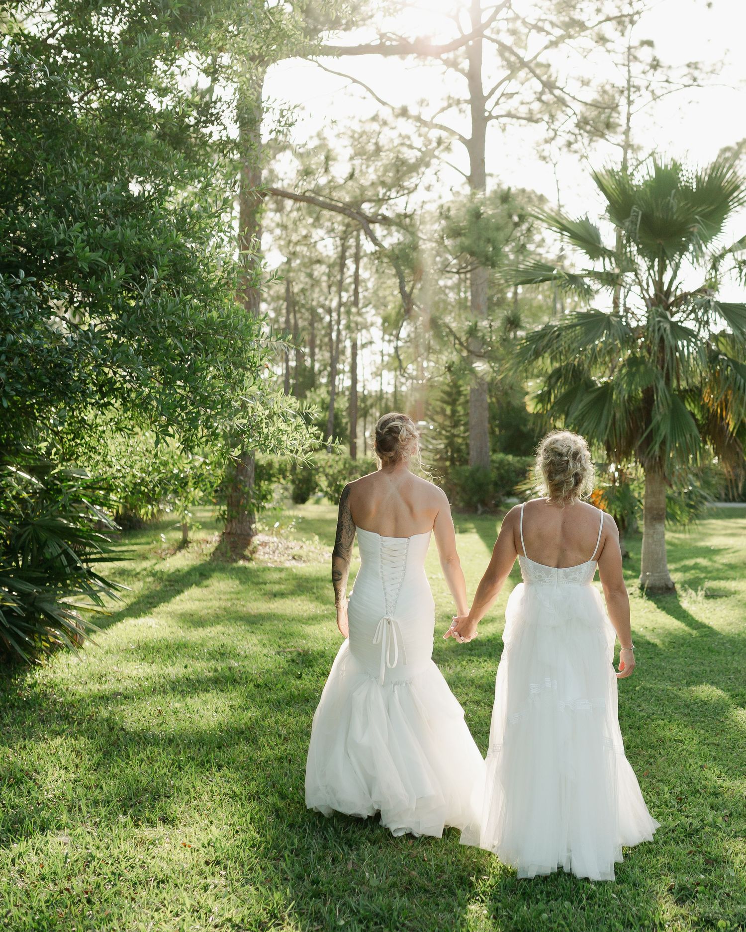 Two brides in white wedding gowns hold hands while walking through a lush green garden setting.