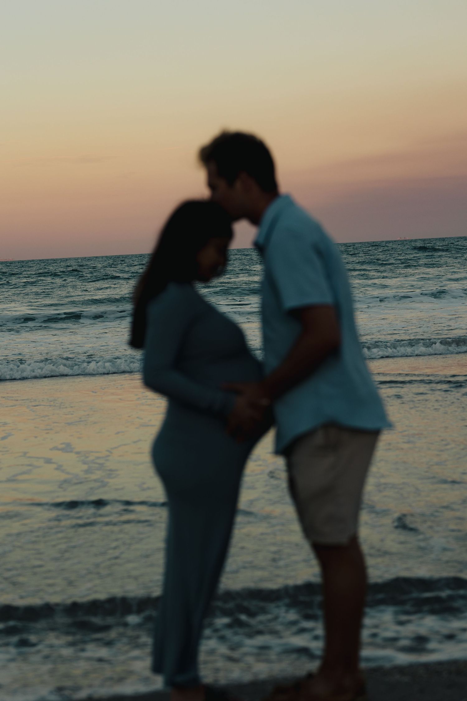 Blurred silhouette of a pregnant couple kissing on the beach during a colorful sunset.