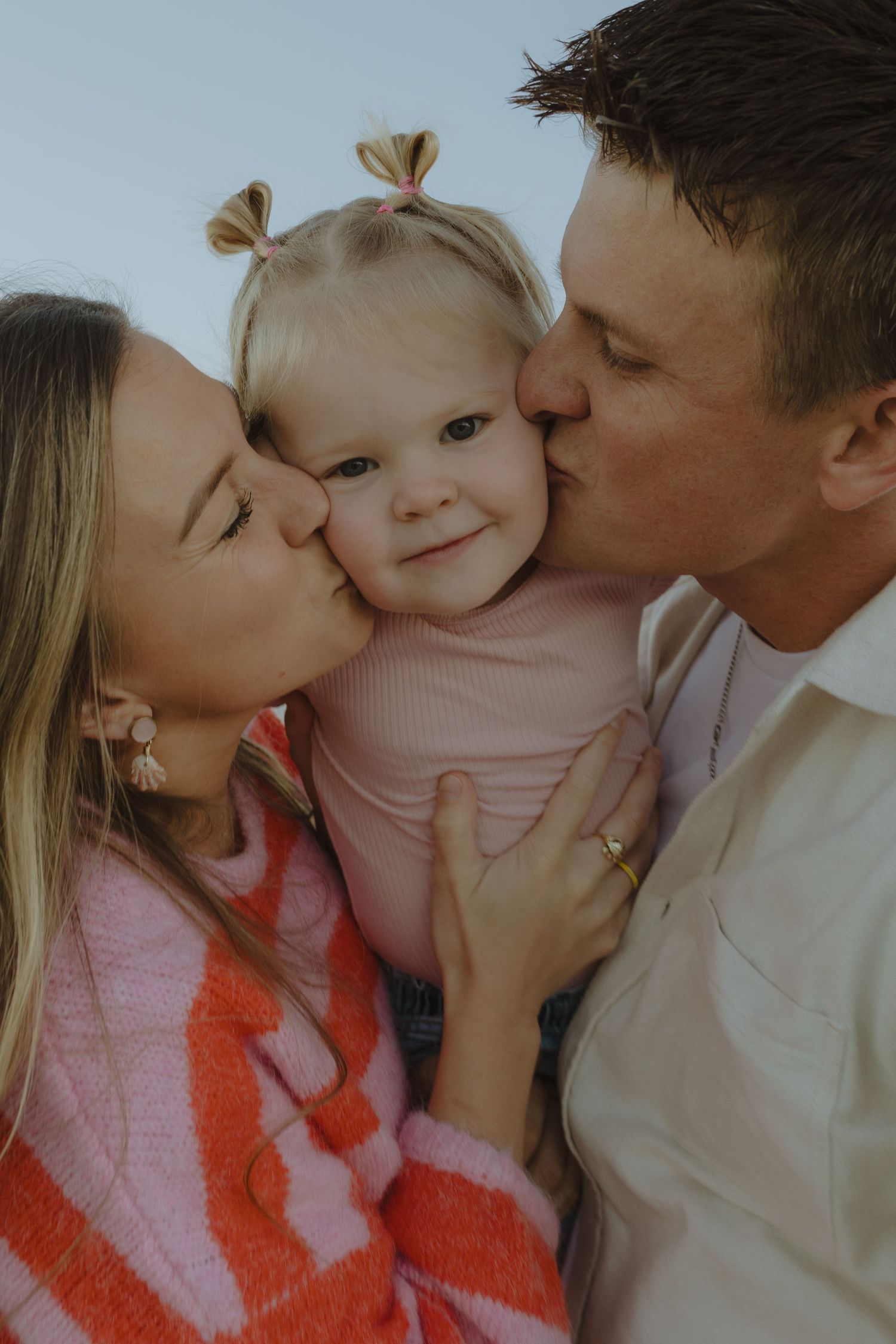 Loving parents simultaneously kissing their calm toddler daughter on the cheeks outdoors.