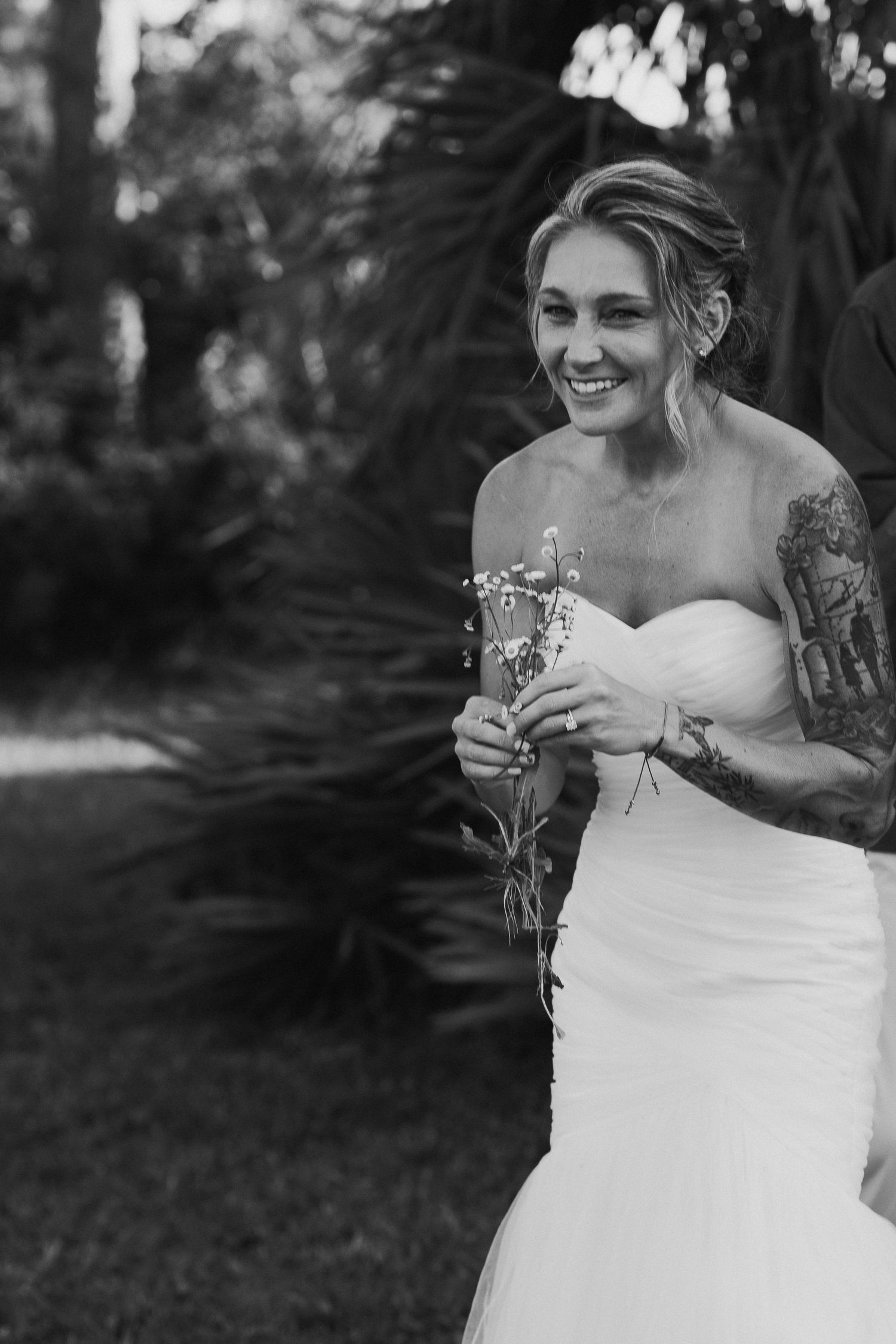 Smiling bride in a strapless white gown with tattoos holds wildflowers in a black and white portrait.