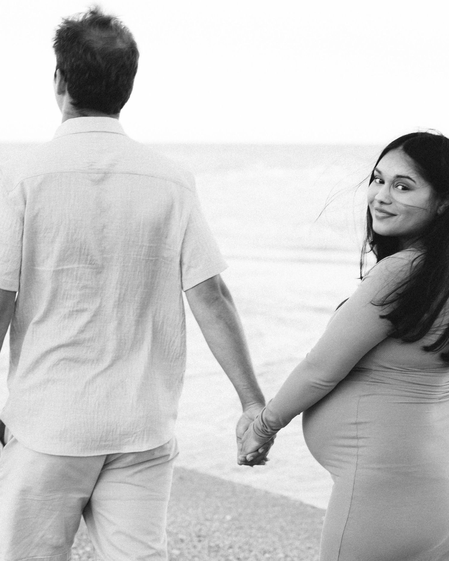 Black and white maternity photo of couple holding hands by the ocean.