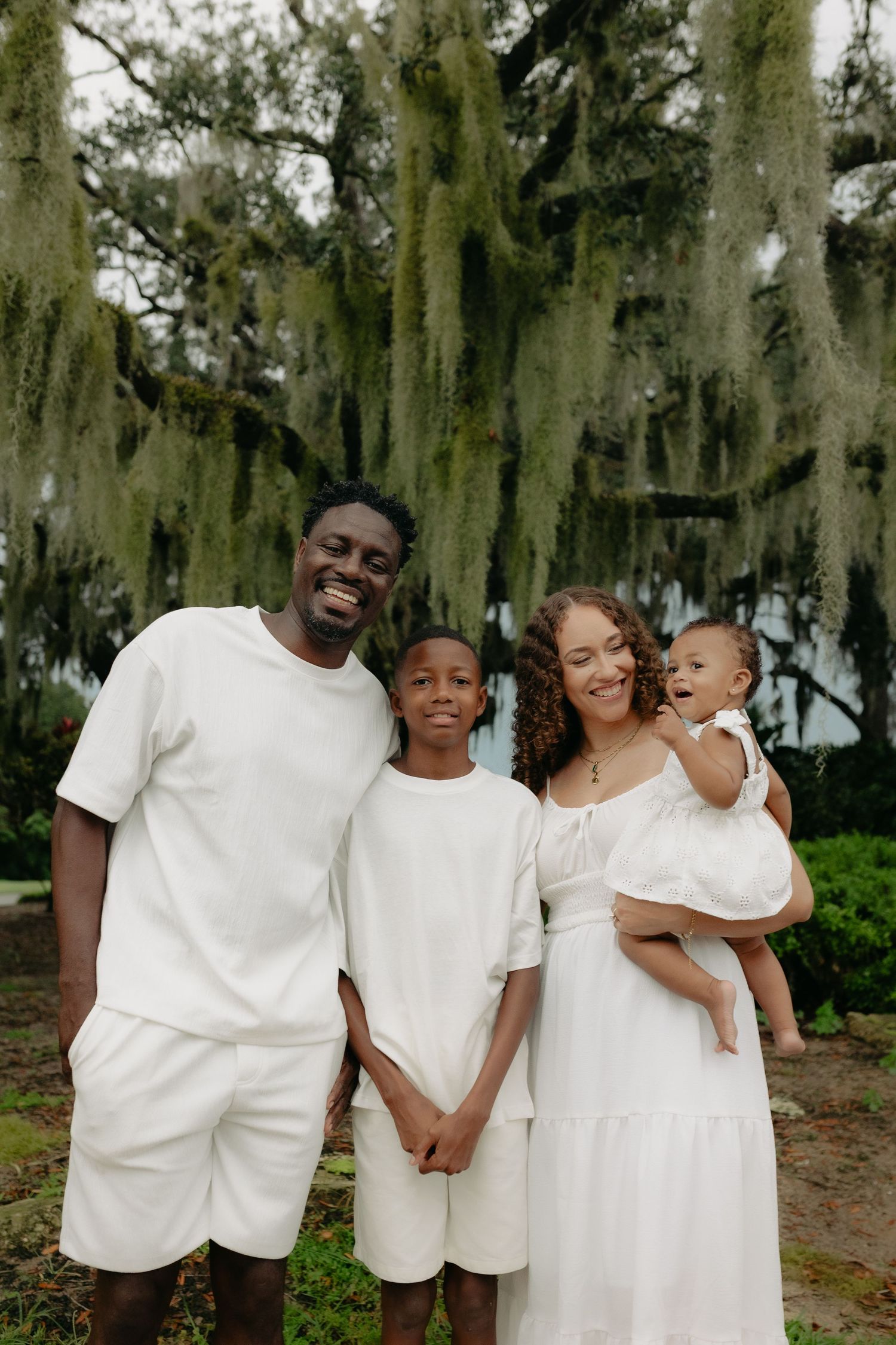 Family of four in white outfits smiling under Spanish moss trees, mother holding baby.