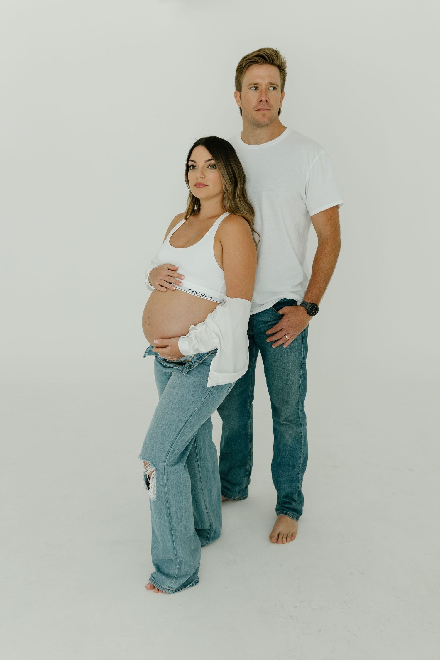 Pregnant couple in matching white tops and jeans posing together in a bright studio setting.