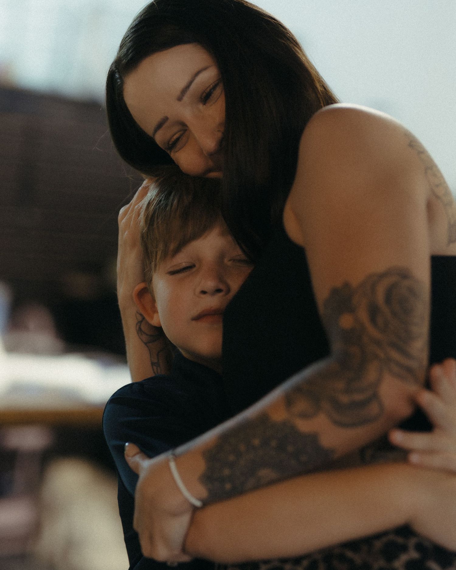 Tattooed mother tenderly kissing her young son on the head while embracing him in a warm hug.