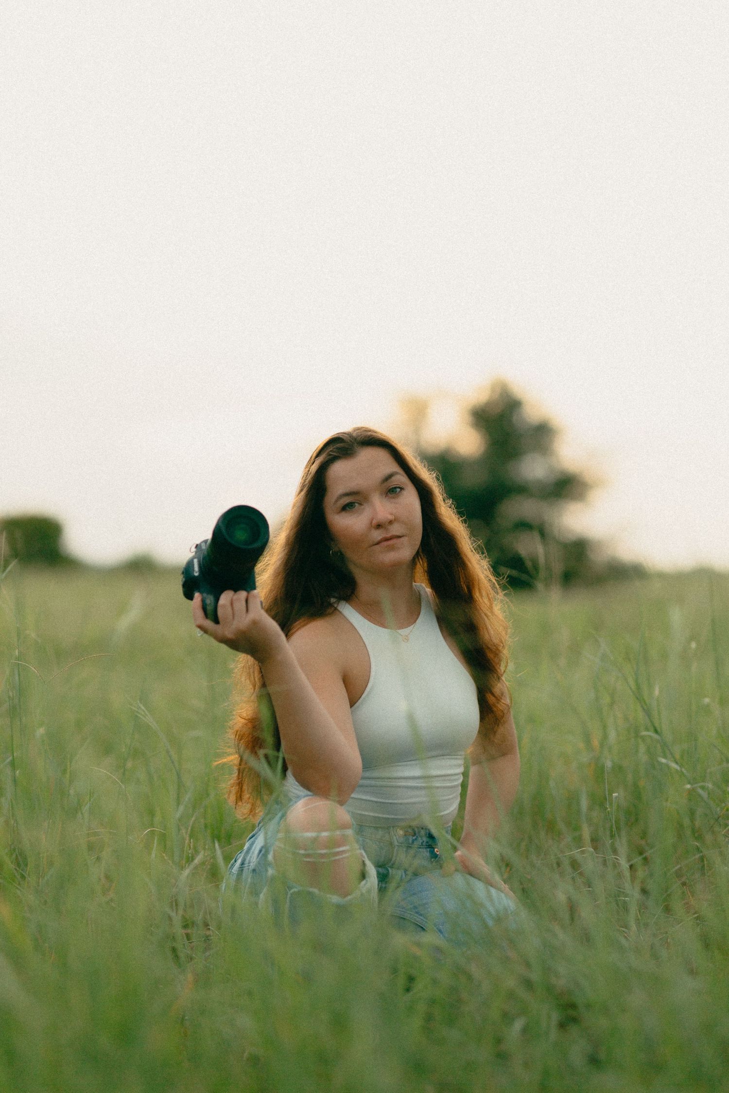 Young woman sitting in tall green grass holding a camera up, wearing a white top and jeans at golden hour.