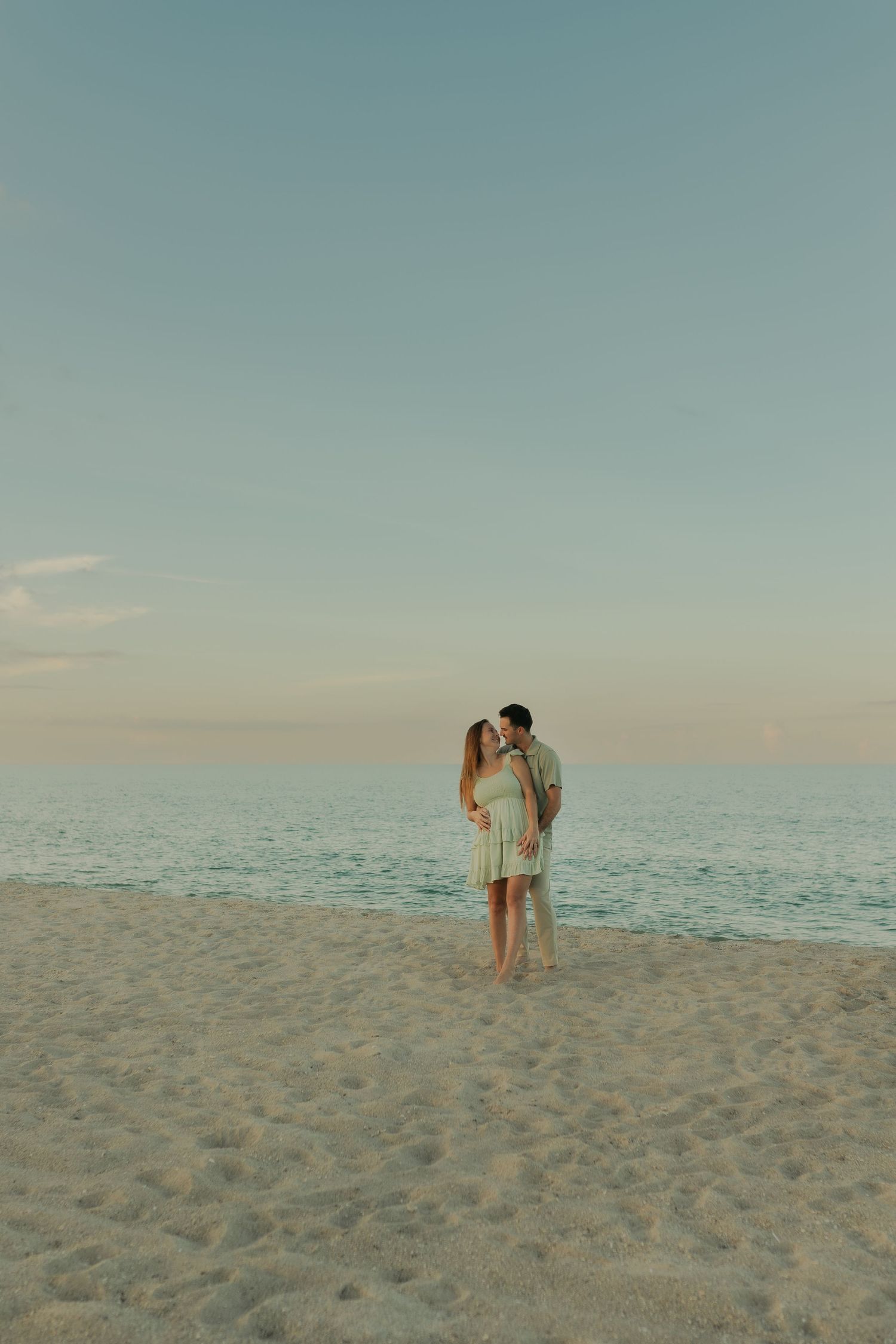 Couple sharing a romantic kiss on a sandy beach at sunset with calm ocean waters in the background.