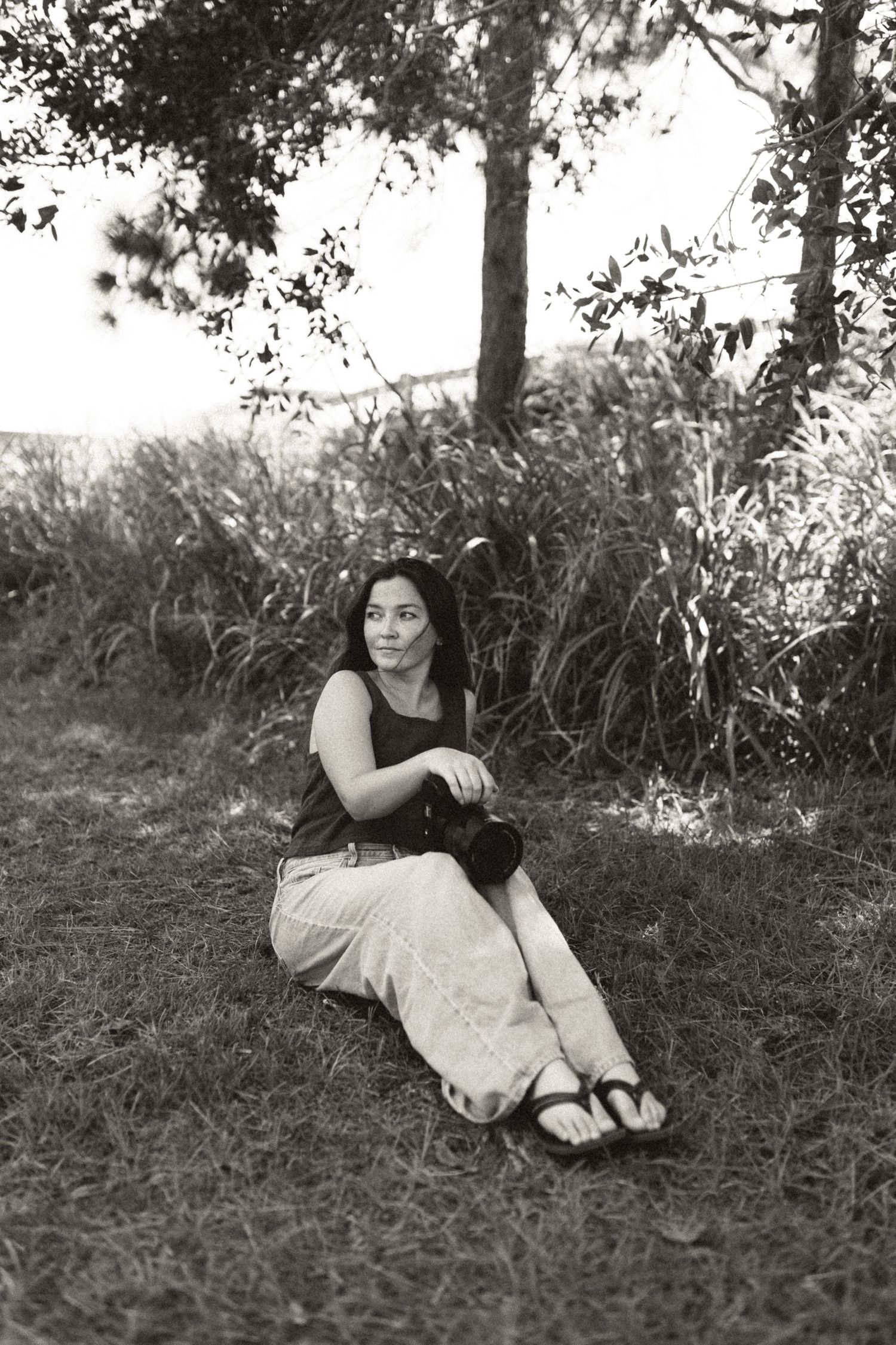 Black and white photo of a woman sitting on grass near trees, holding a camera, wearing a tank top and light skirt.