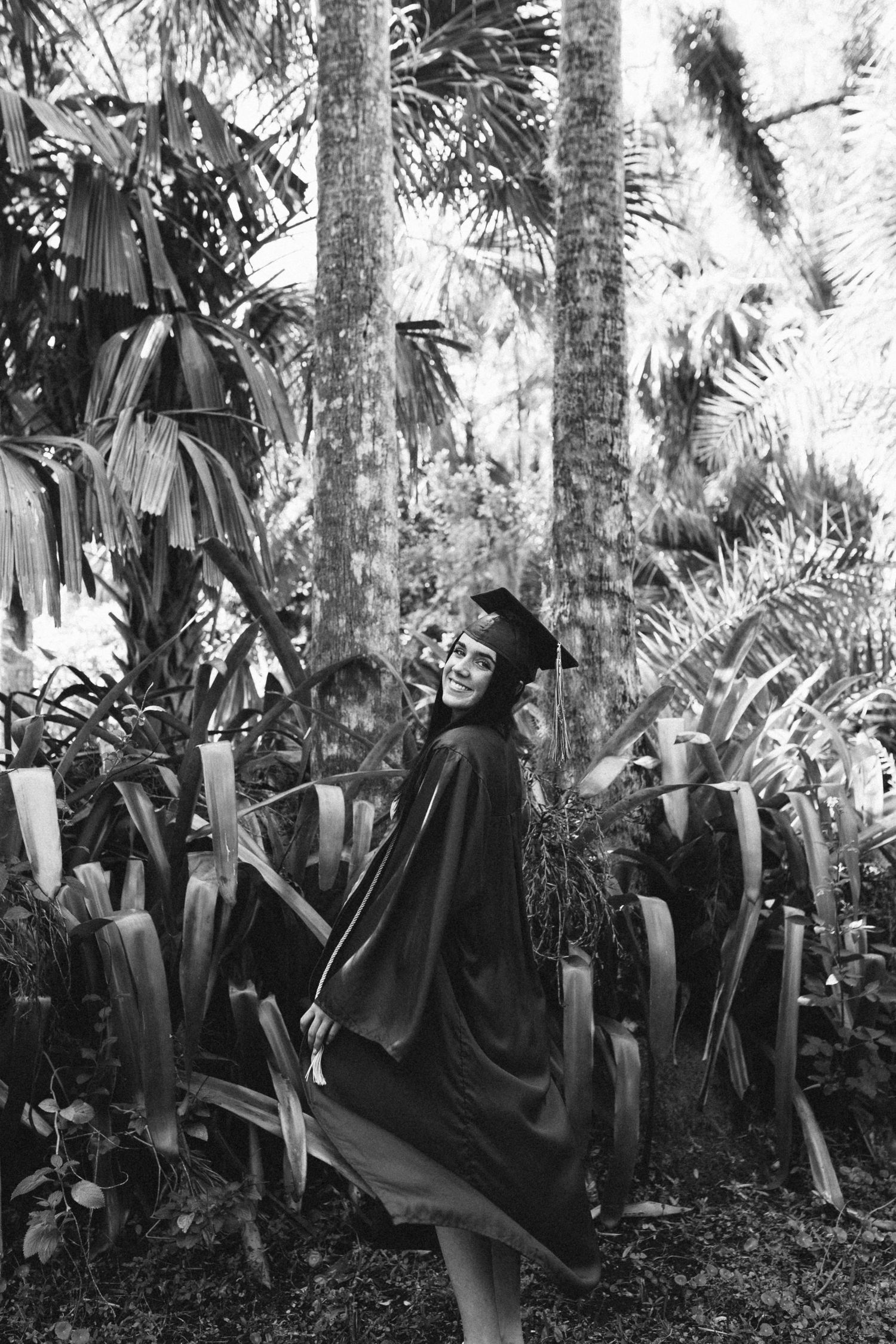 Black and white photo of a woman in graduation cap and gown posing among lush tropical foliage.