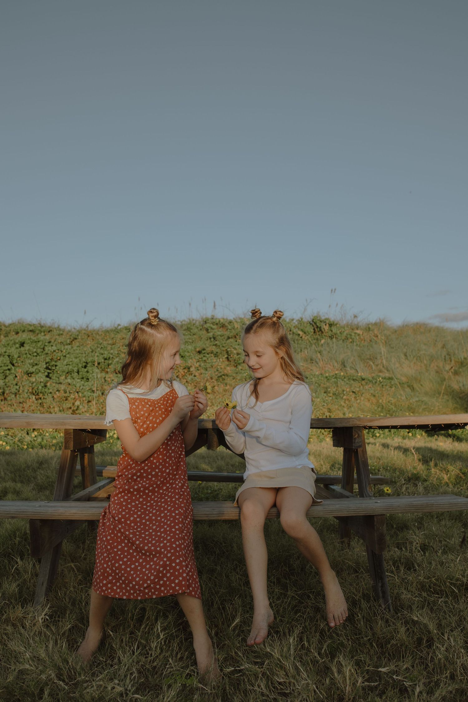 Two young girls in summer outfits sitting on wooden bench outdoors sharing small flowers at sunset.