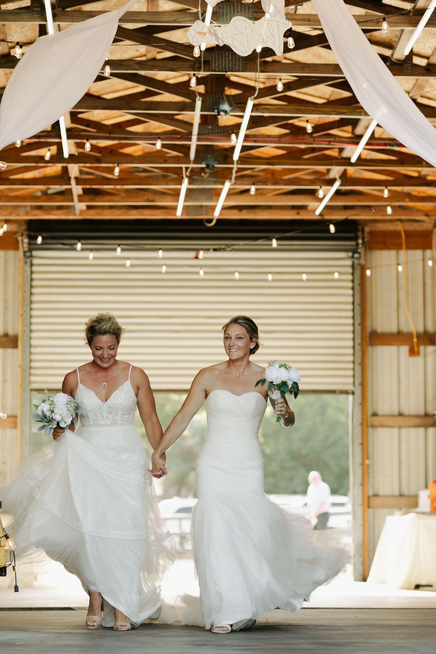 Two brides in white wedding gowns hold hands and smile while walking through a decorated barn venue.