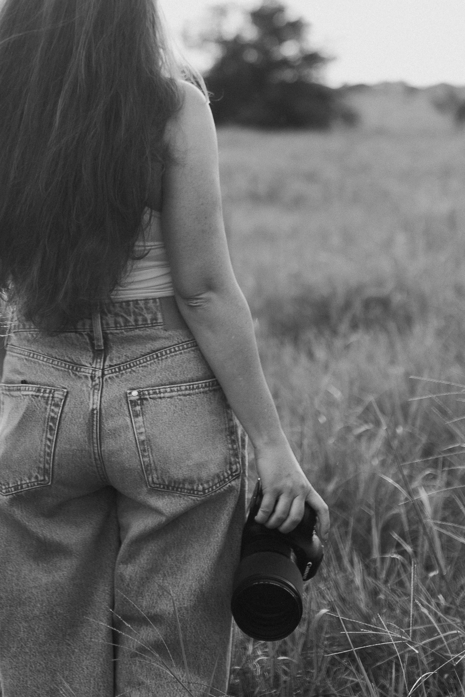 Black and white rear view of female photographer in jeans holding camera while walking through field.