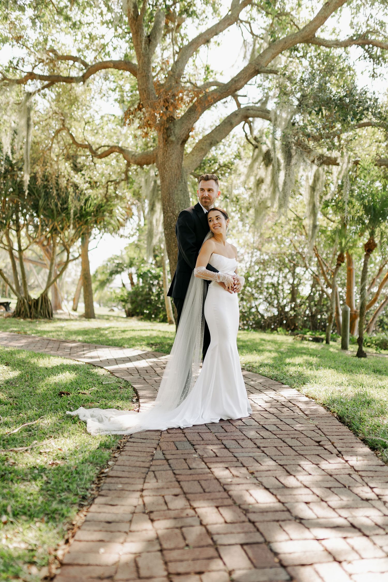 Bride and groom pose on a brick path beneath sprawling oak trees draped with Spanish moss.