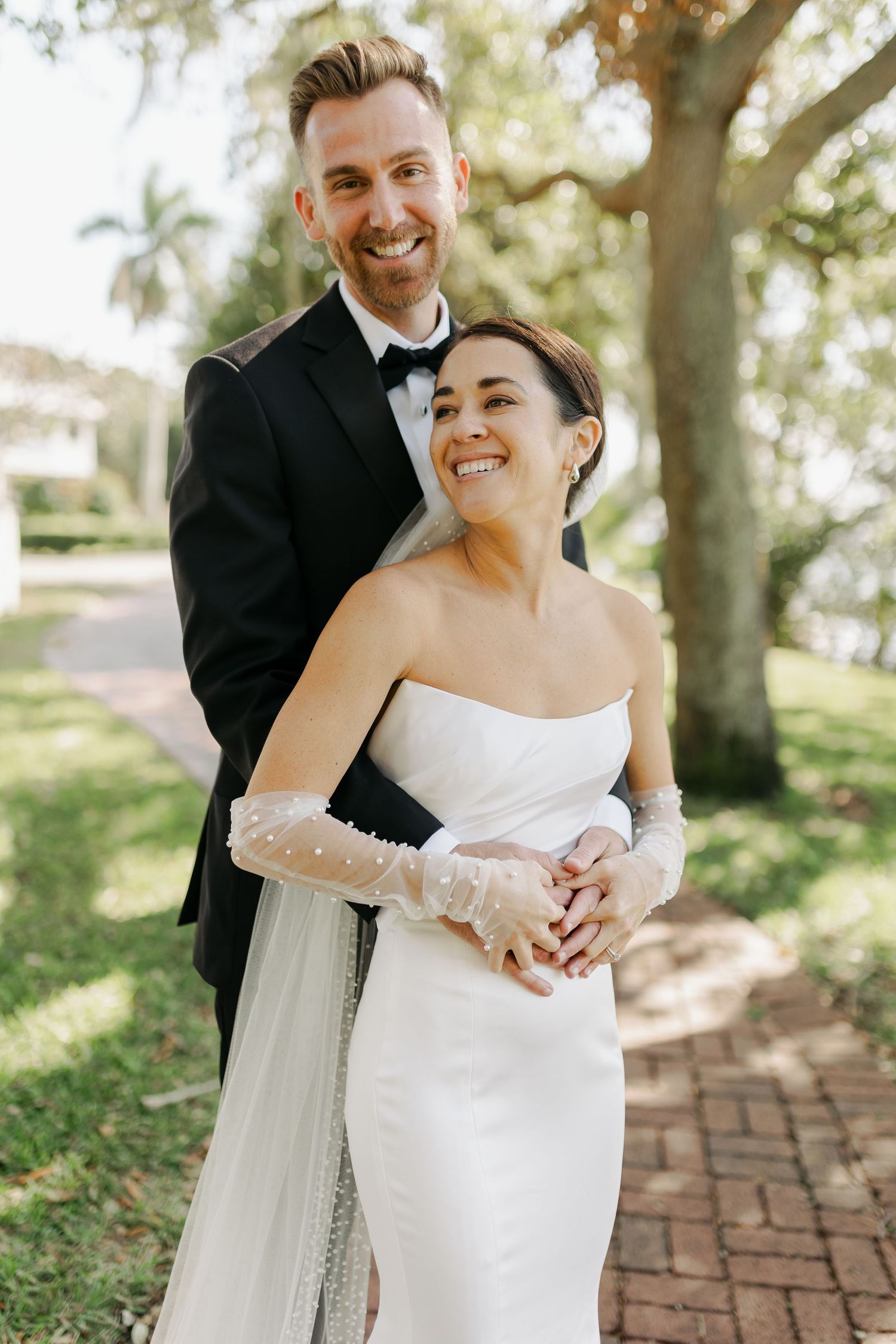 Smiling bride and groom embracing outdoors in elegant wedding attire under lush green trees.