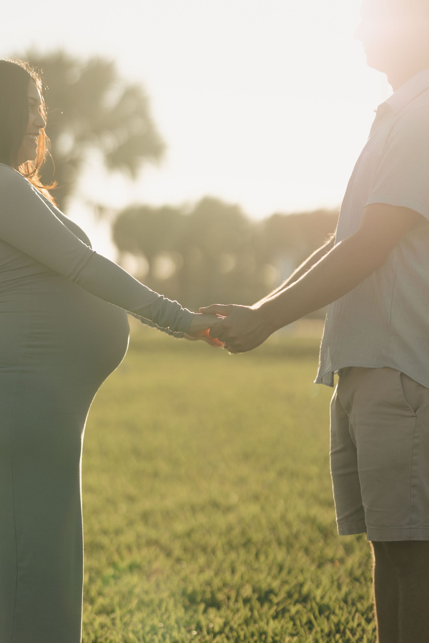 Pregnant couple holding hands in a sunlit green field during golden hour at sunset.