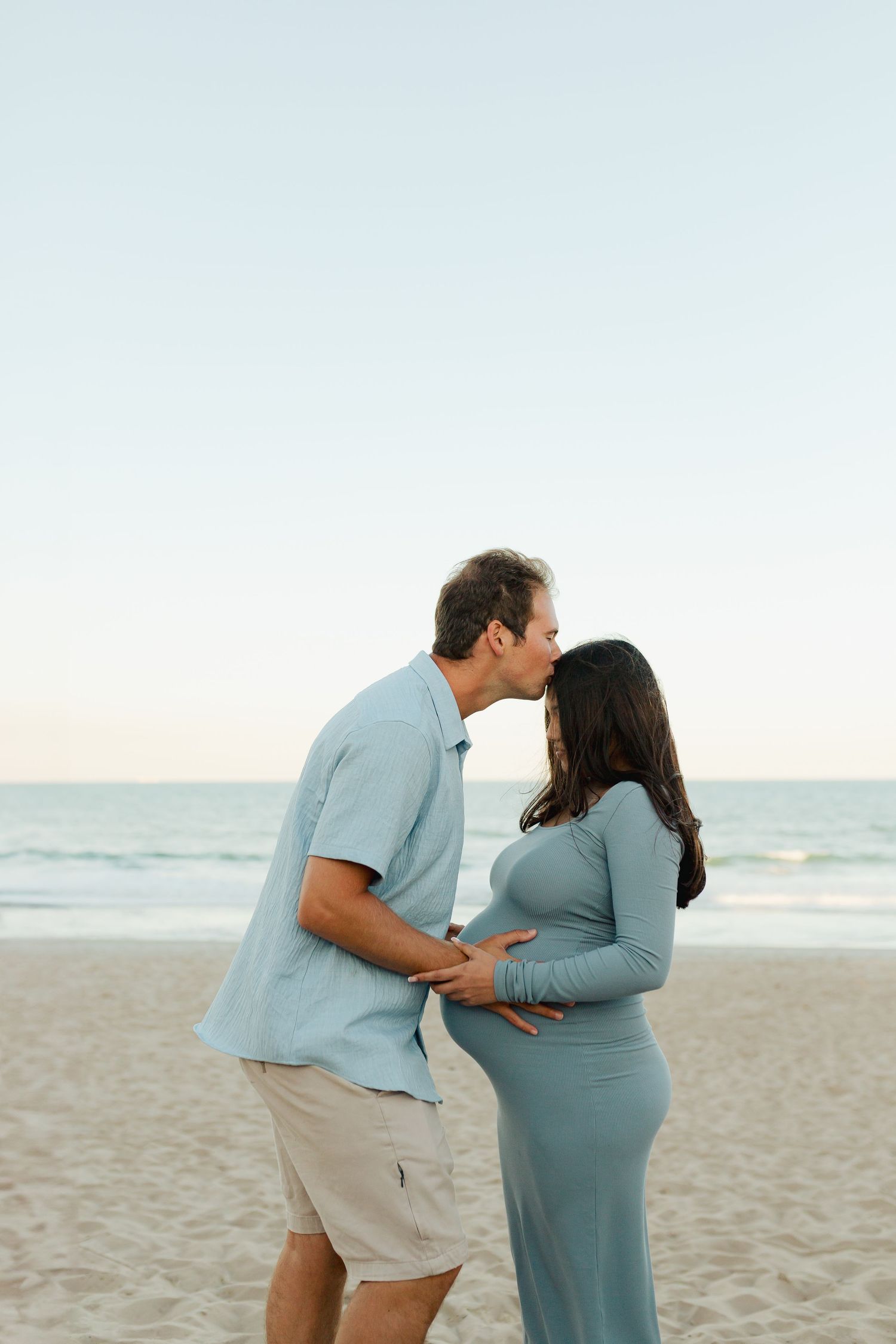 Couple sharing a kiss on the beach, man gently holding pregnant woman's baby bump at sunset.