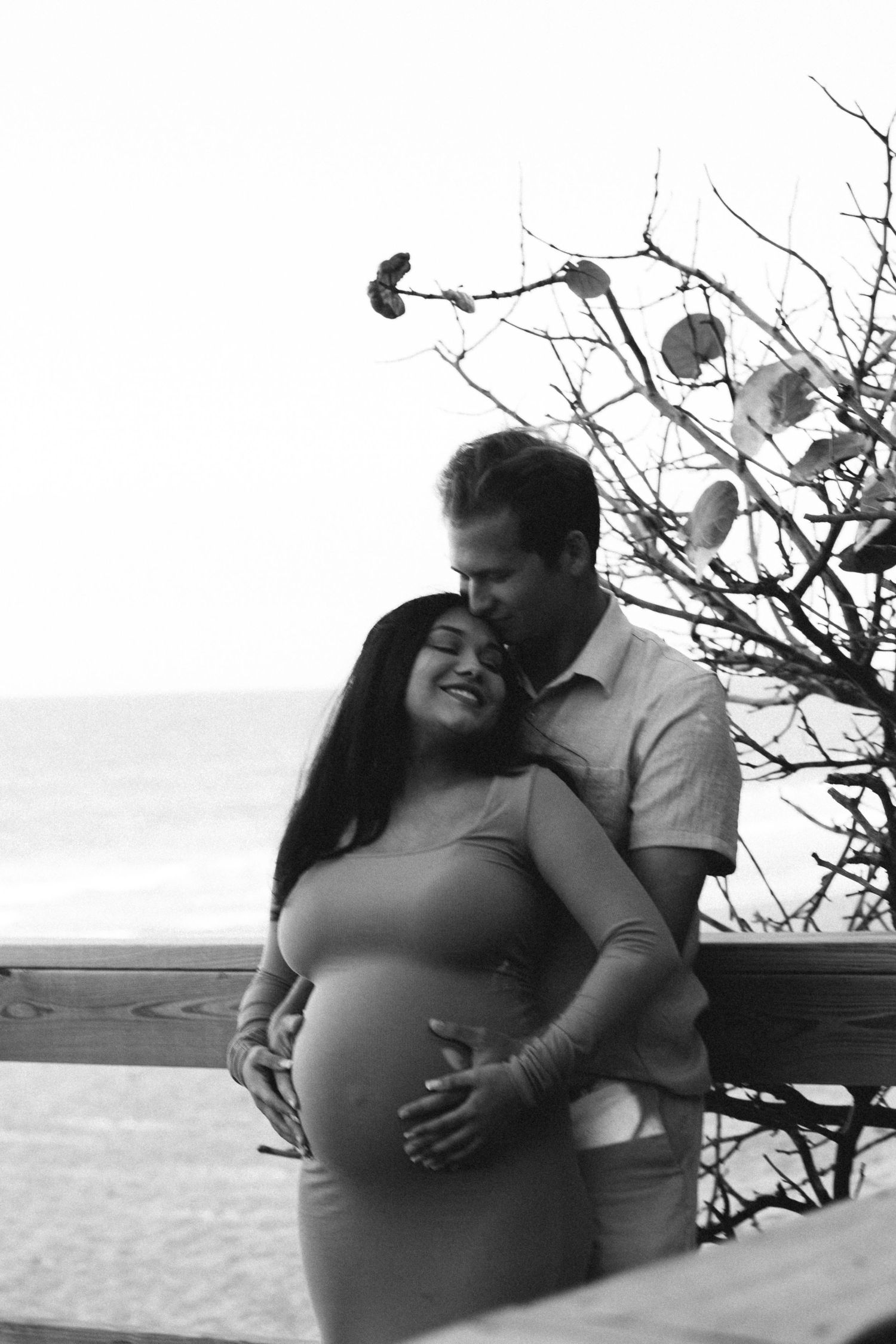 Black and white photo of a couple embracing outdoors near the ocean, man kissing pregnant woman.