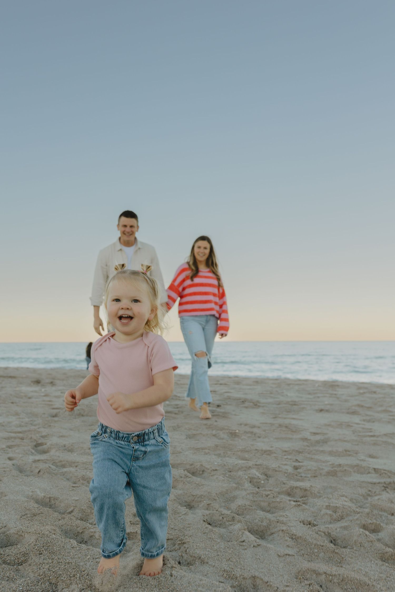 Happy toddler running on sandy beach toward camera with parents smiling in background at sunset.
