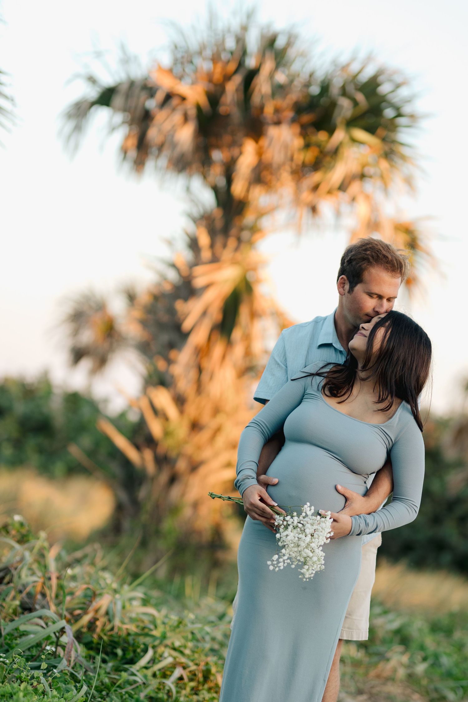 Couple embracing near a palm tree at golden hour, man kissing pregnant woman holding white flowers.