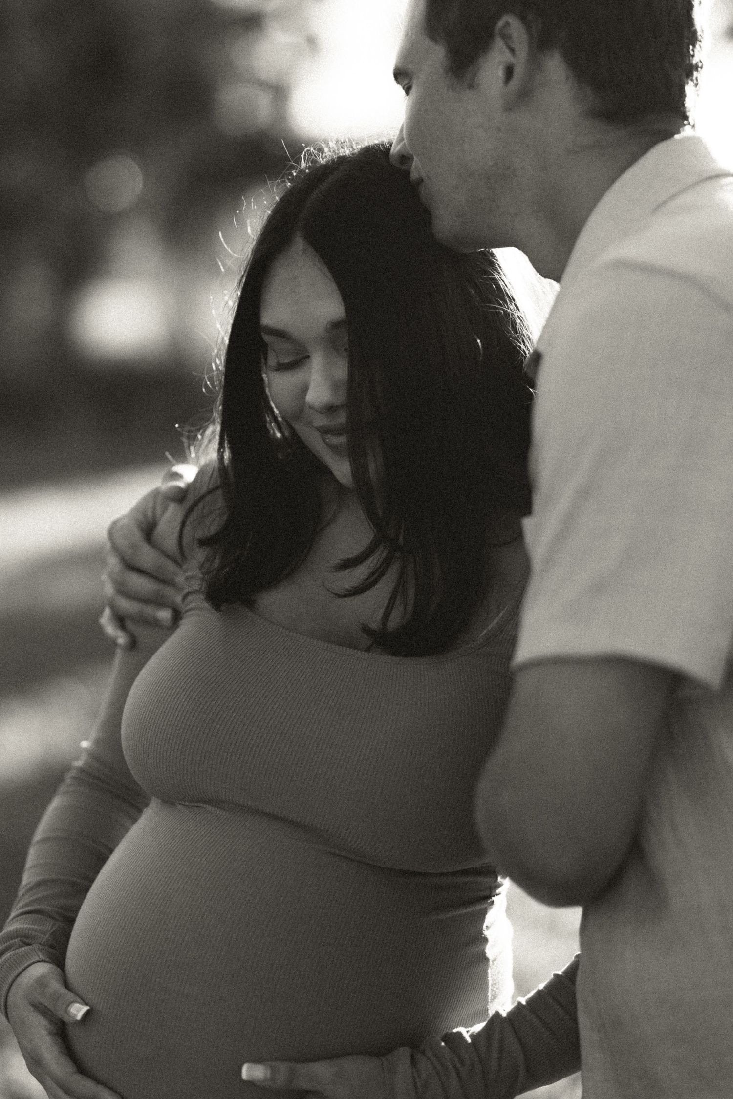 Black and white photo of a man kissing a smiling pregnant womans head while she holds her bump.